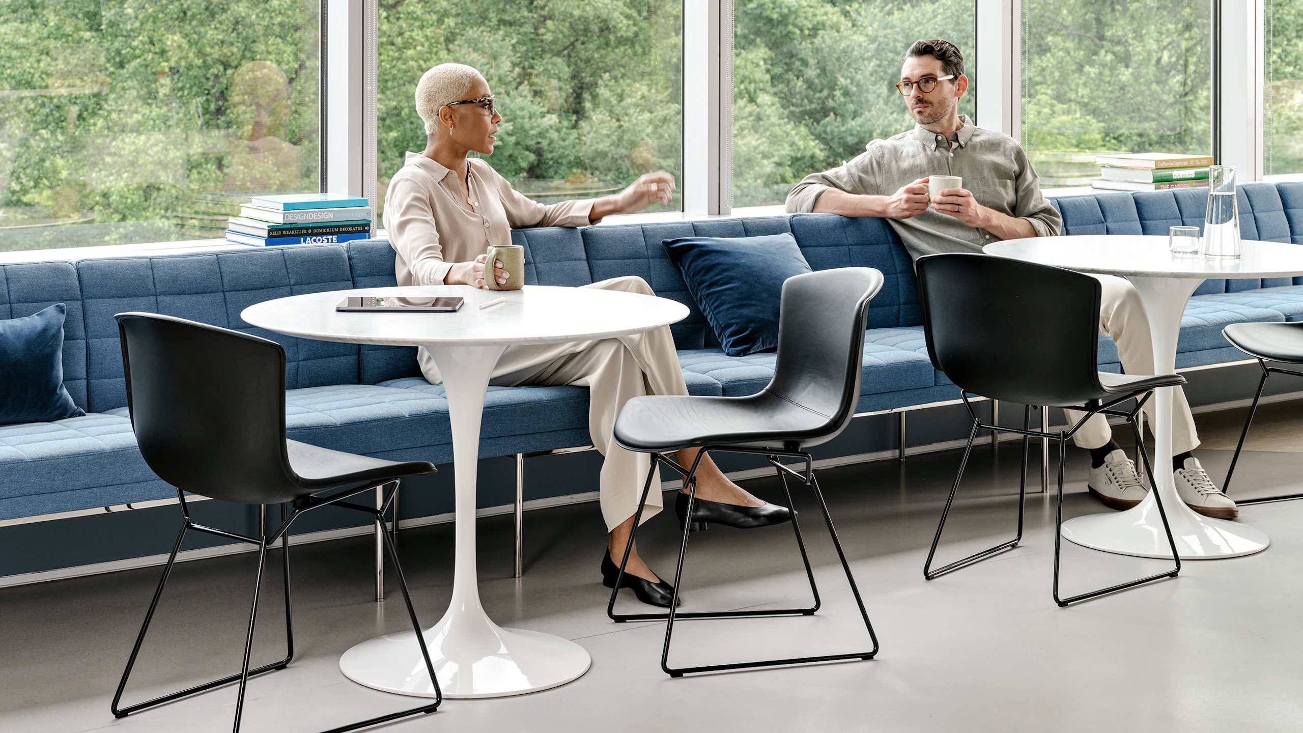 Two individuals conversing in a shared seating area furnished with Saarinen Tables, Knoll Bertoia chairs, and blue bench seating