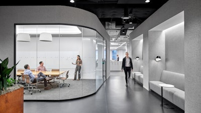 Três pessoas usando uma grande sala de conferências com janelas abertas, mobiliada com cadeiras de alumínio Eames da Herman Miller, enquanto um indivíduo passa por uma fileira de sofás cinza e mesas laterais brancas