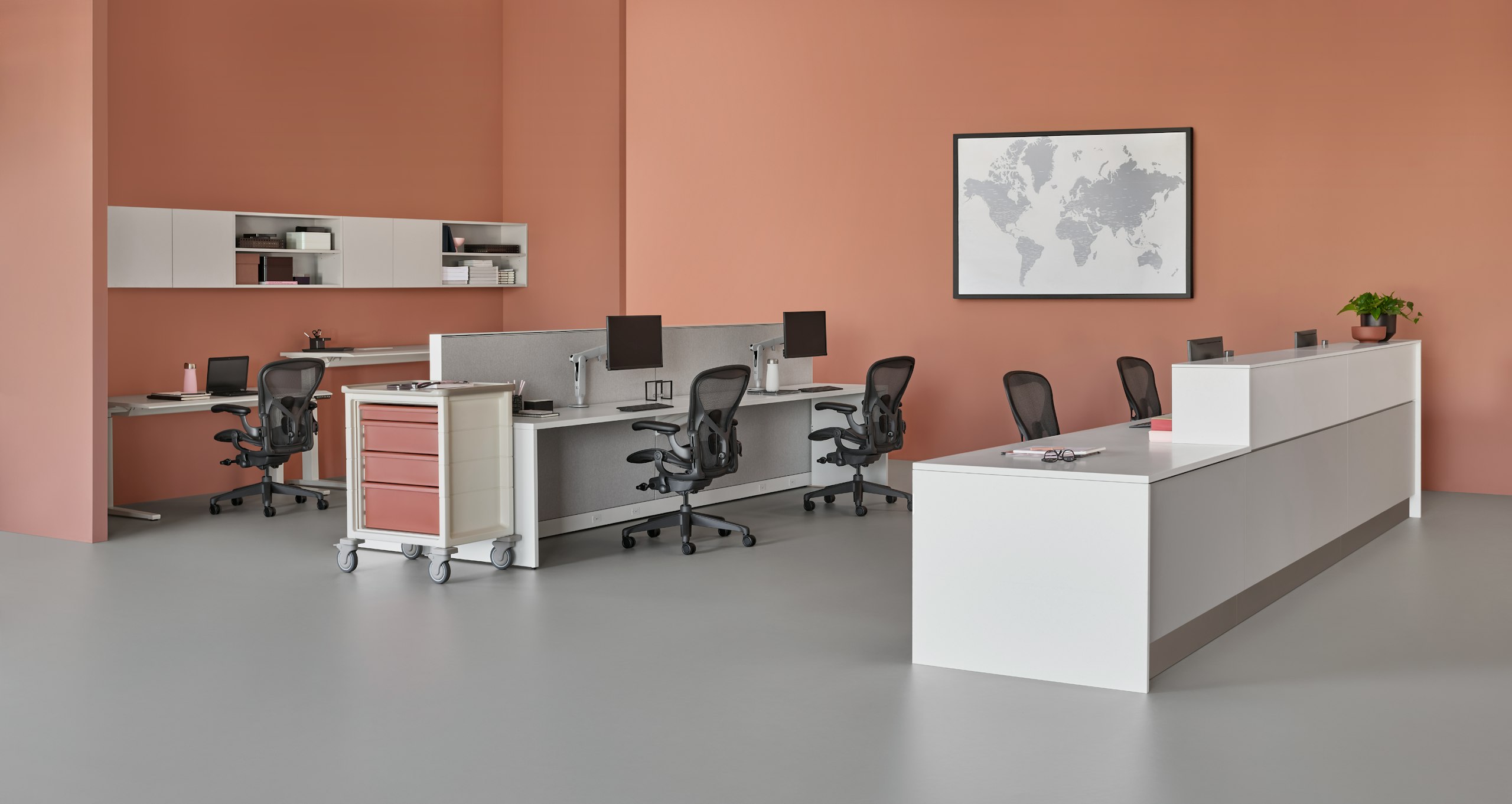 A caregiver environment setting with a white and gray prefab Commend Nurses Station, Renew Sit-to-Stand Tables, and Aeron Chairs.