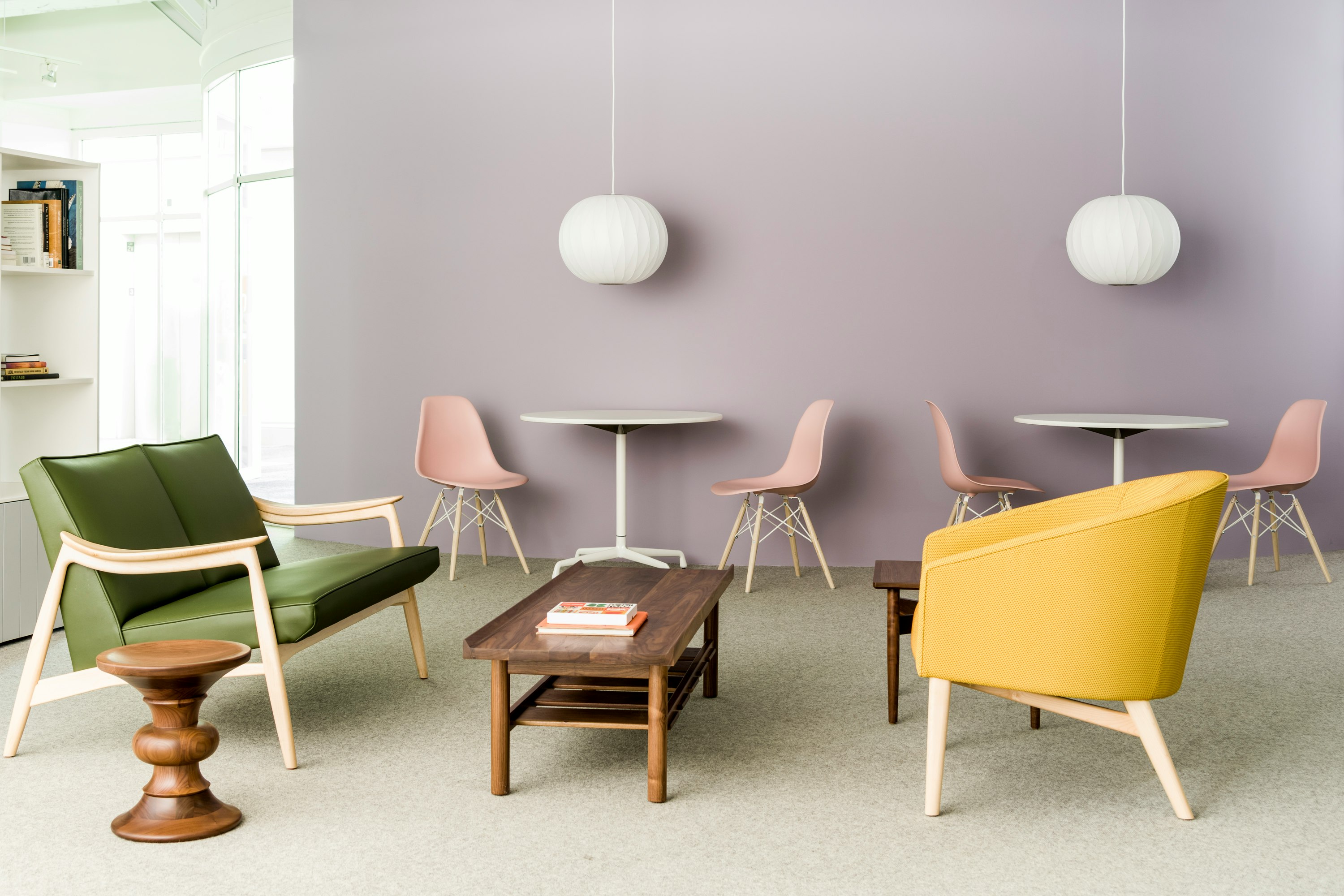 A brightly lit environment featuring a green Aspen Settee, dark wood Tamarack Table and Bench, and mustard yellow Sophora Lounge Chair, with blush pink Eames shell chairs in the background.