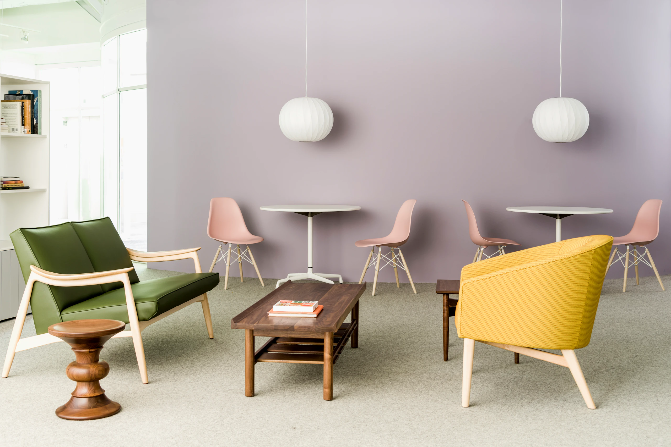 A brightly lit environment featuring a green Aspen Settee, dark wood Tamarack Table and Bench, and mustard yellow Sophora Lounge Chair, with blush pink Eames shell chairs in the background.
