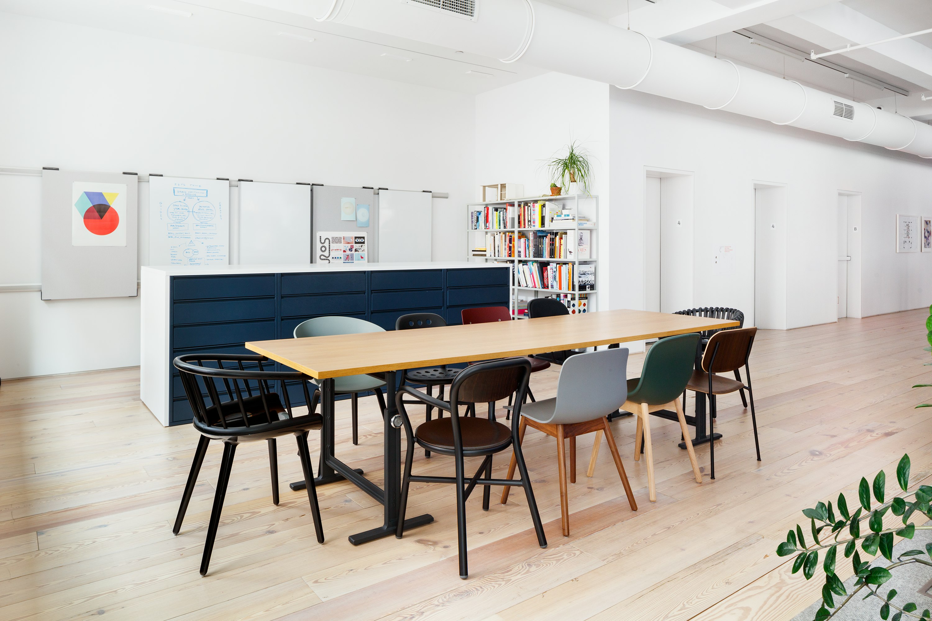Angled view of light brown Brut Table, with black legs, surrounded by medley of chairs in various colors.