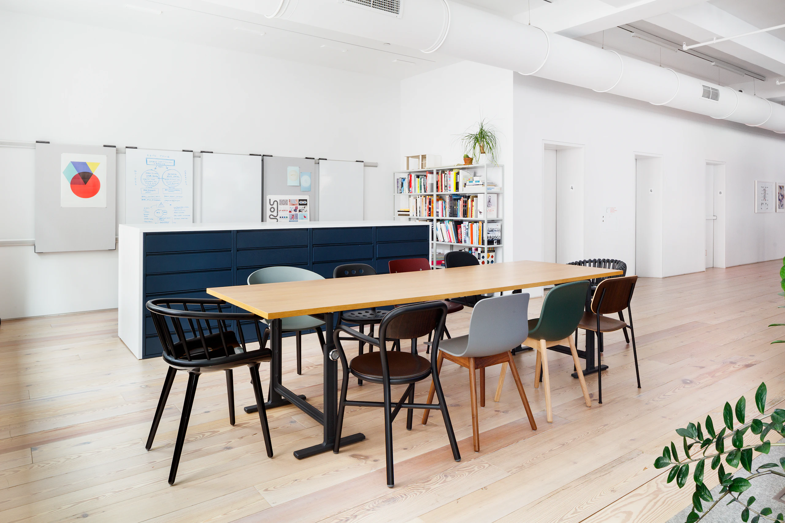 Angled view of light brown Brut Table, with black legs, surrounded by medley of chairs in various colors.