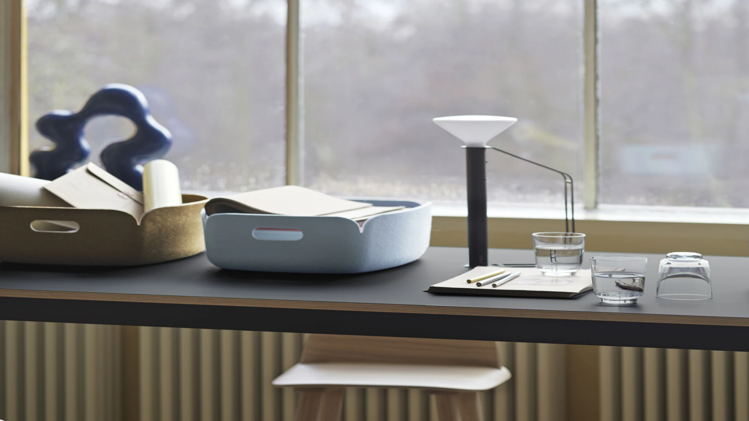 Two Restore Baskets by Muuto, one blue and one yellow, atop a counter height table next to a notepad, lamp, and glasses of water. Sunlight floods through a large window.