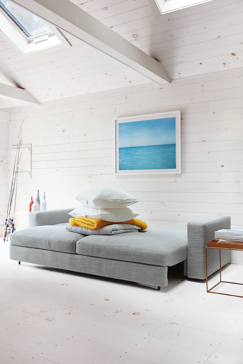 Bright living room scene featuring a gray sectional sofa, stacked cushions, a yellow throw, and a framed ocean artwork on a white wooden wall.