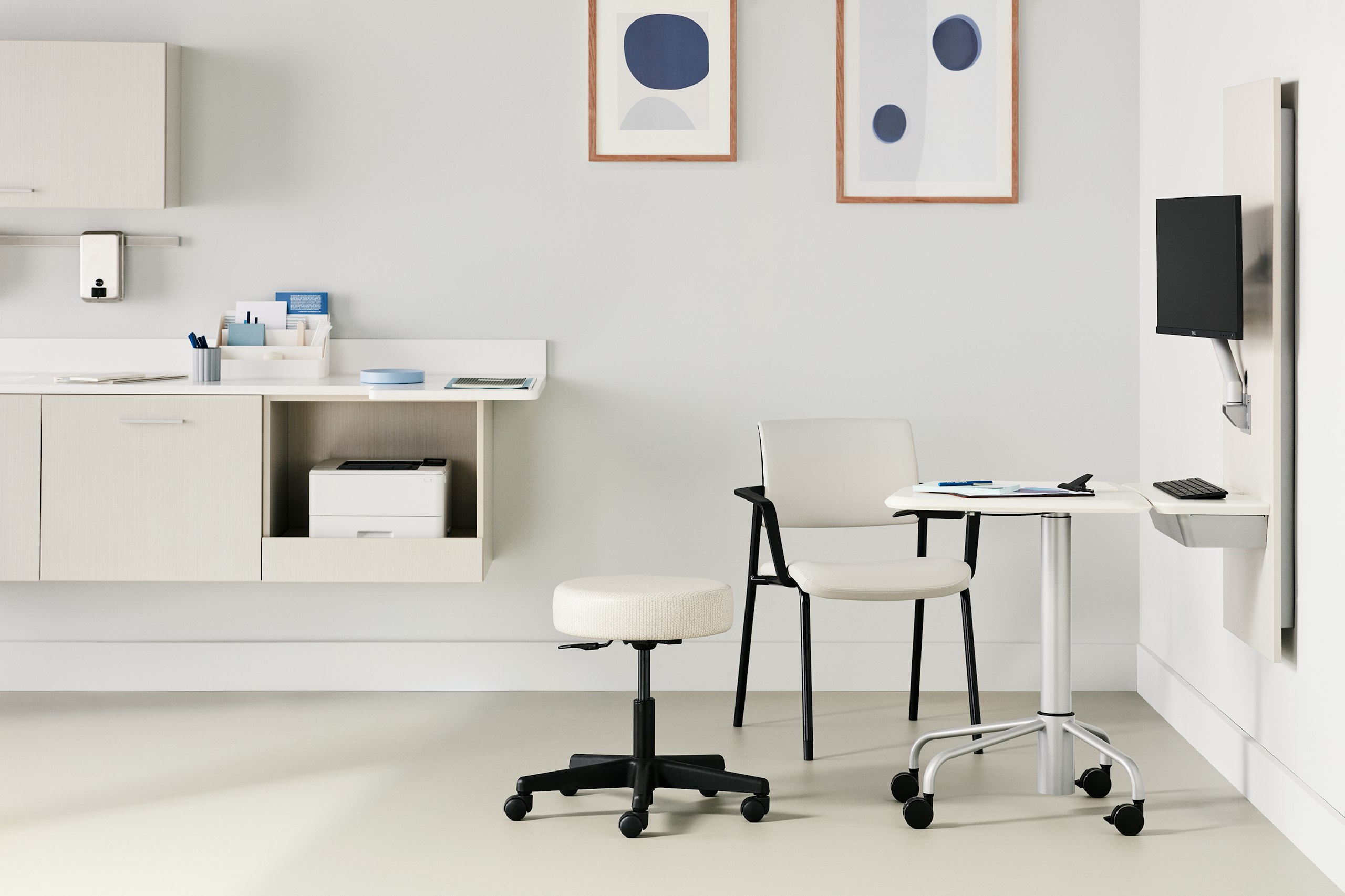 Exam room with a Mora casework unit in a light twill finish mounted on the back wall and an Intent wall unit in a light twill finish mounted on the side wall. There's a mobile, height-adjustable table nested to the wall unit with a white top and silver metal legs. A side chair fully upholstered in a light gray upholstery and a black frame. A physician's stool in a light gray upholstery and a black base.