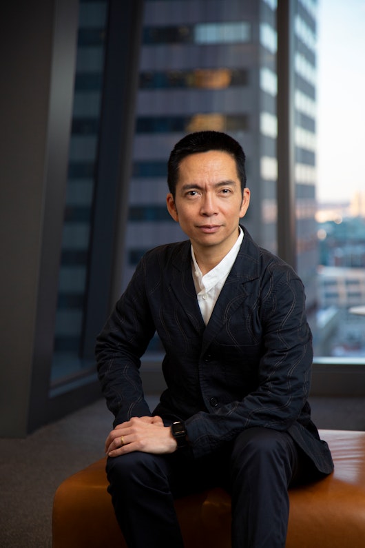 John Maeda, a member of the Board of Directors of MillerKnoll, Inc., wearing a dark blue suit while seated in front of a large window overlooking a city