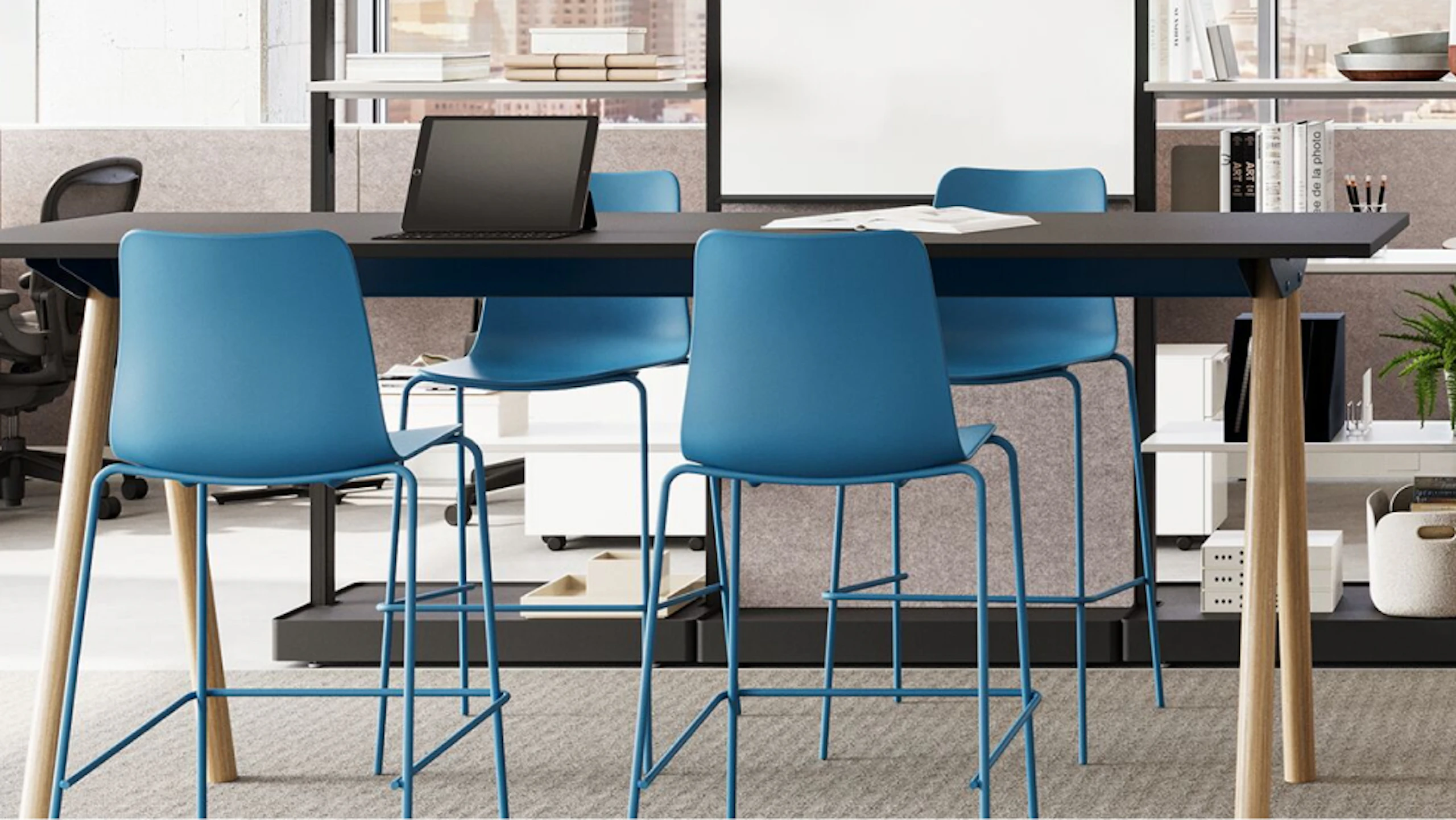 A black NaughtOne Morse Bar Table with wooden legs surrounded by four blue bar-height chairs in a team neighbourhood setting designed to facilitate change, reduce disruption and improve resiliency.