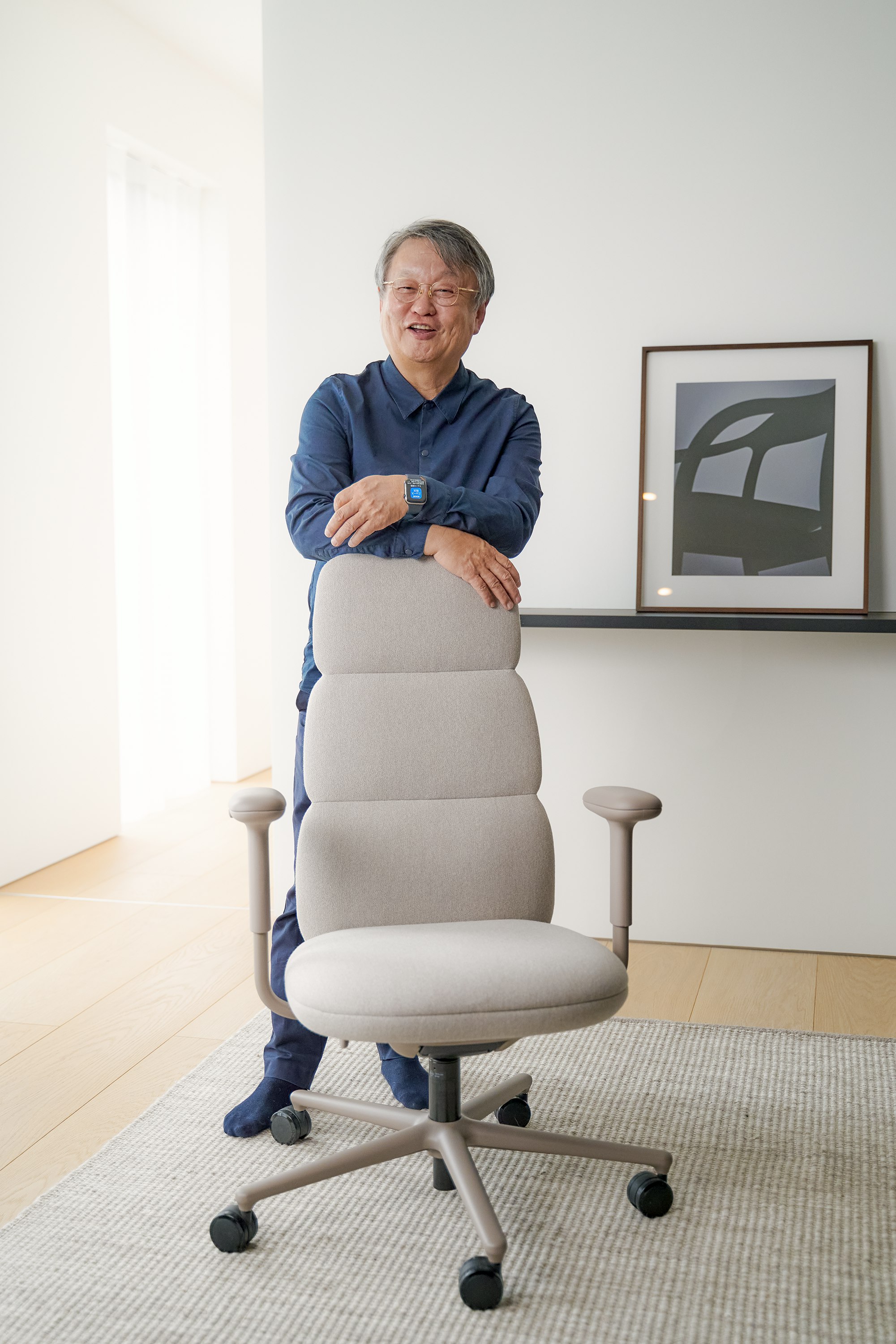 Designer Naoto Fukasawa standing behind a high back Asari chair in light brown.
