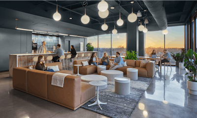 Different groups of people interacting in a communal space that features brown sofas, bar seating, green potted plants, and floor to ceiling windows
