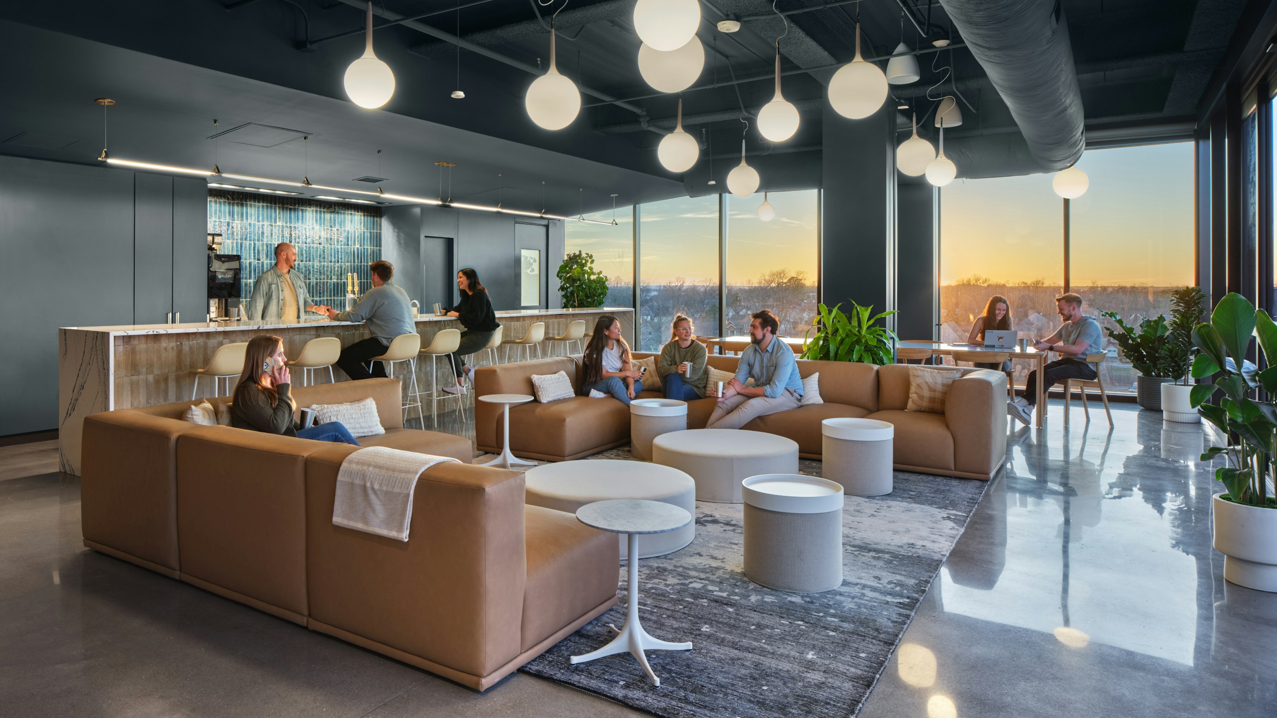 Different groups of people interacting in a communal space that features brown sofas, bar seating, green potted plants, and floor to ceiling windows