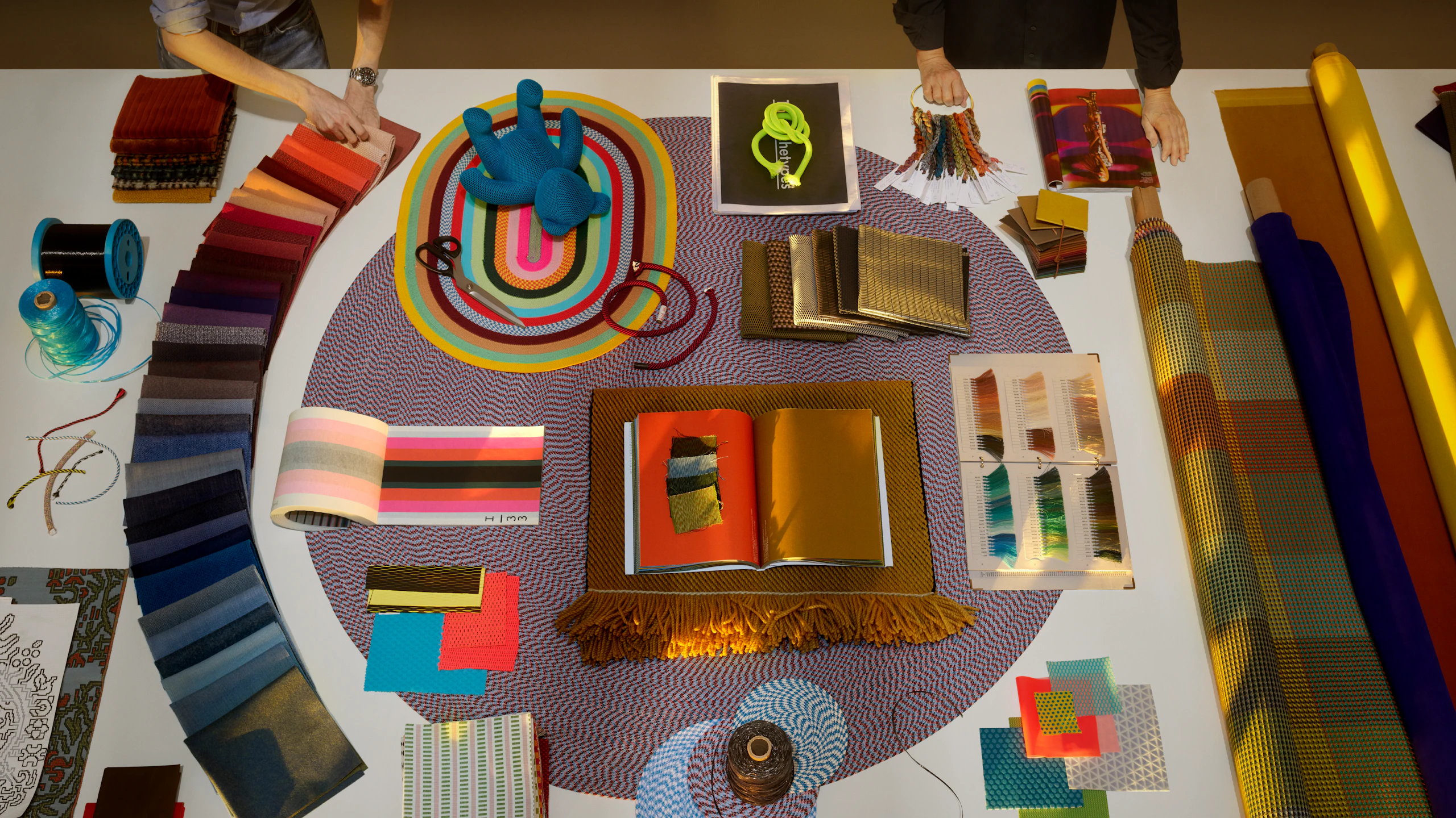 Two individuals interacting with a colorful assortment of Maharam textiles laid out on a white table.
