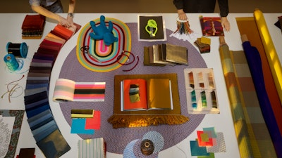 Two individuals interacting with a colorful assortment of Maharam textiles laid out on a white table.