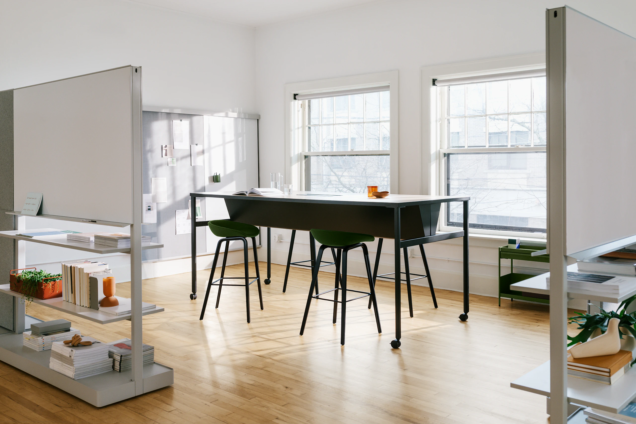 A black double sided OE1 Communal Table with casters, with OE1 Agile Walls in the foreground and OE1 Wall Rail and Board and OE1 Storage Trolley in the background.