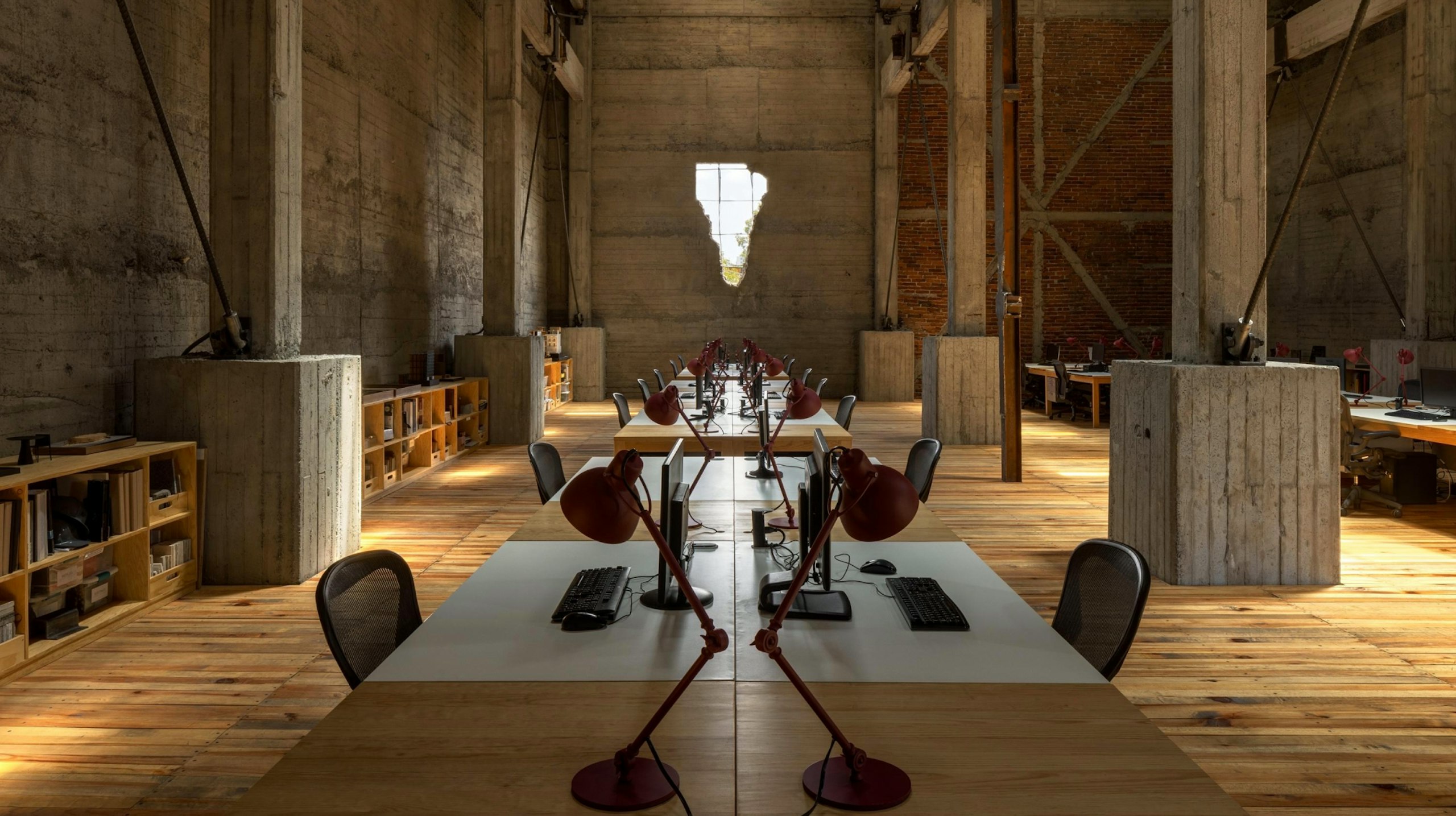 Rows of workstations with computers, red lamps and task chairs at the JSa office in an old industrial building with large concrete beams