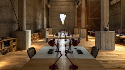 Rows of workstations with computers, red lamps, and task chairs at the JSa office in an old industrial building with large concrete beams