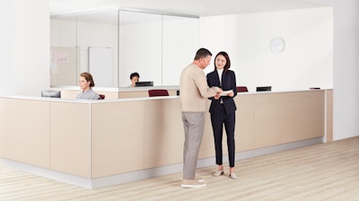 A physician speaking with a patient in front of a prefab, light ash woodgrain Commend Nurses Station with two nurses working at the desk.