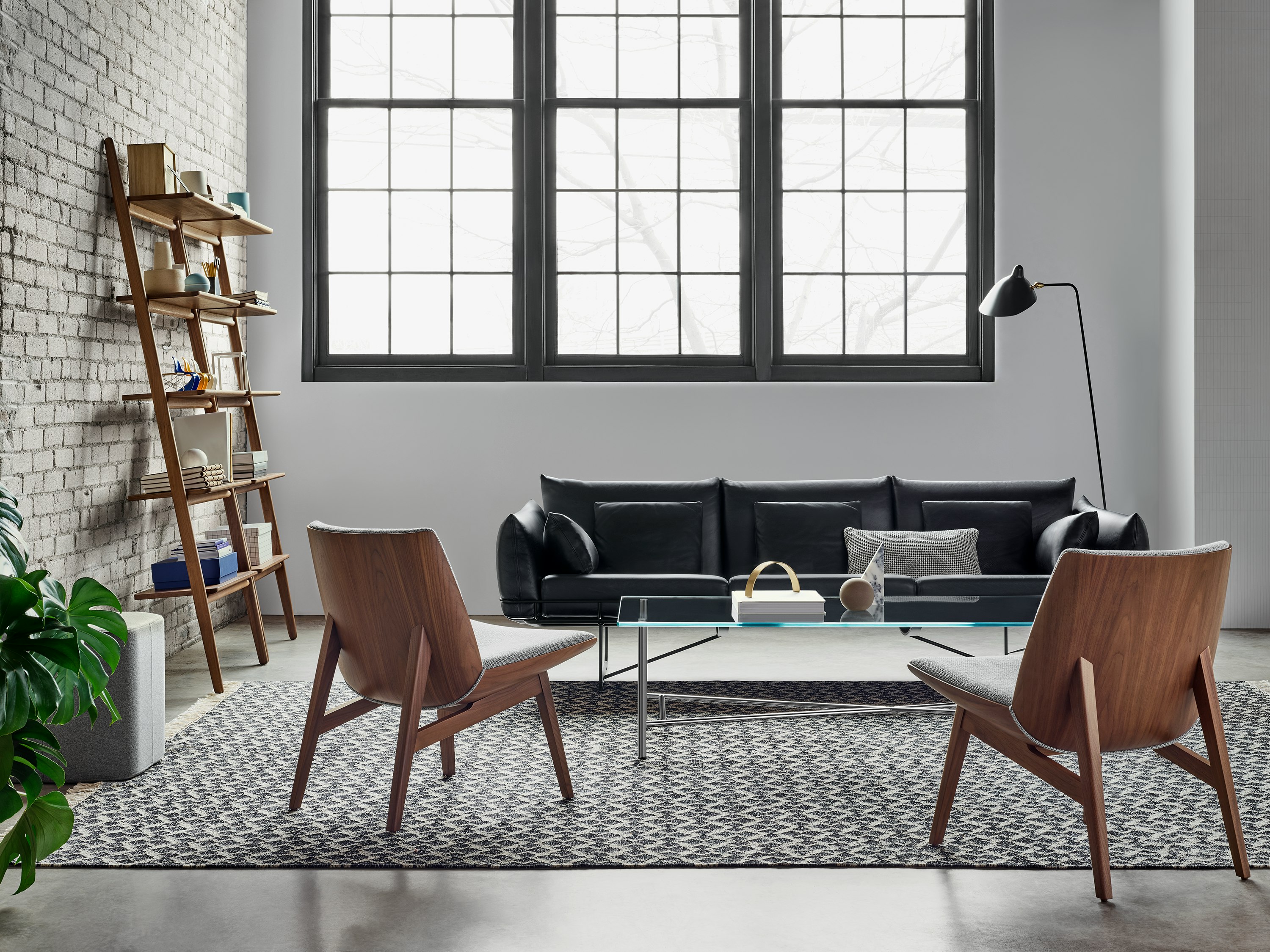 Two low back Clamshell Lounge Chairs in a light grey textile and walnut base as well as a black Wireframe Sofa are around a glass Layer Table. The scene is set on a patterned rug in front of a brick wall with windows, a shelving unit, and a black Mantis BS1 B Floor Lamp.