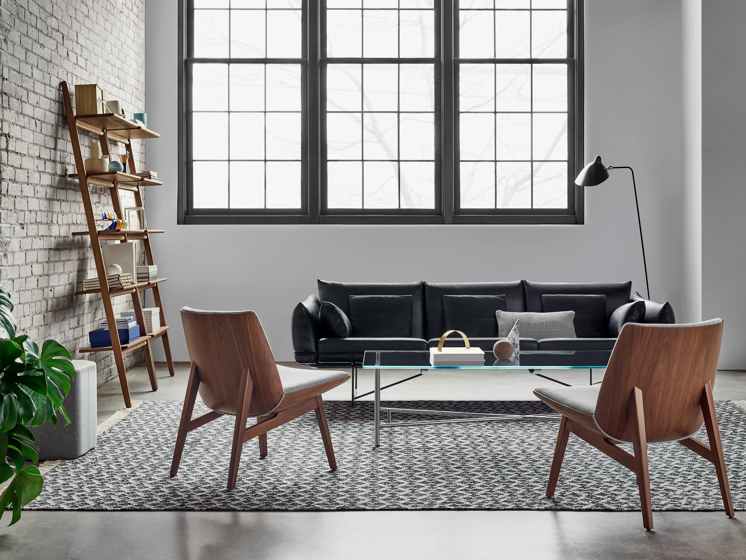 Two low back Clamshell Lounge Chairs in a light grey textile and walnut base as well as a black Wireframe Sofa are around a glass Layer Table. The scene is set on a patterned rug in front of a brick wall with windows, a shelving unit, and a black Mantis BS1 B Floor Lamp.