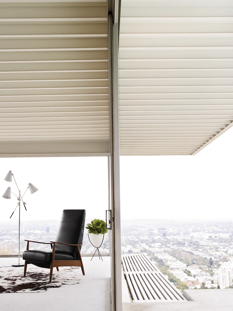 Apartment balcony scene with mid-century chair inside a glass-walled living area; cityscape in background.