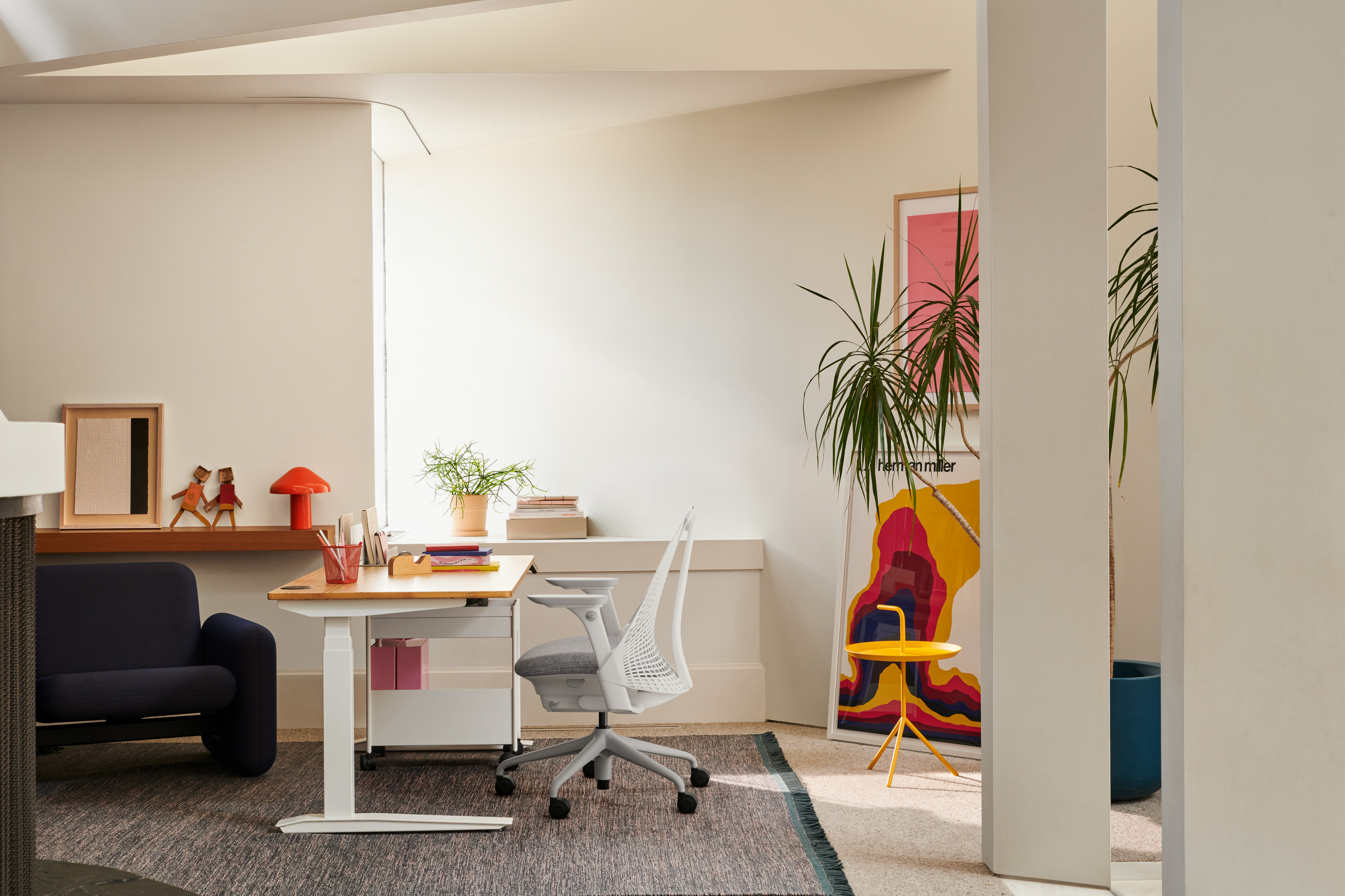 Side view of a bright office featuring a white Herman Miller Sayl Office Chair, Jarvis Bamboo Sit-Stand Desk, Wilkes Modular Armchair, HAY Pao Portable Lamp and OE1 Storage Trolley.