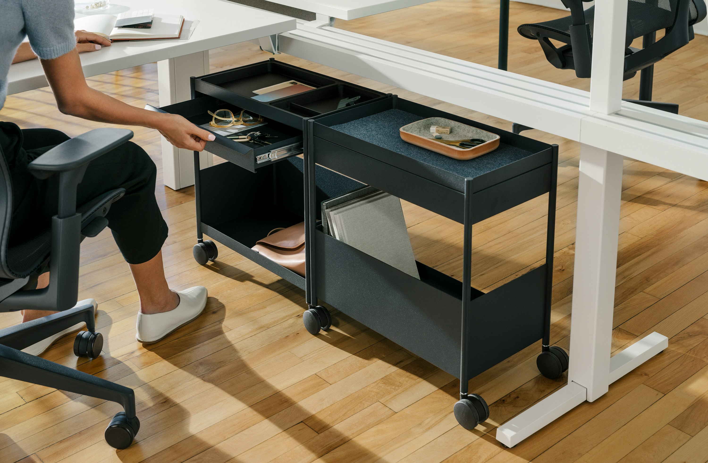  A woman opens a drawer on a blue, shared OE1 Storage Trolley that nests under a white Canvas Vista chase.