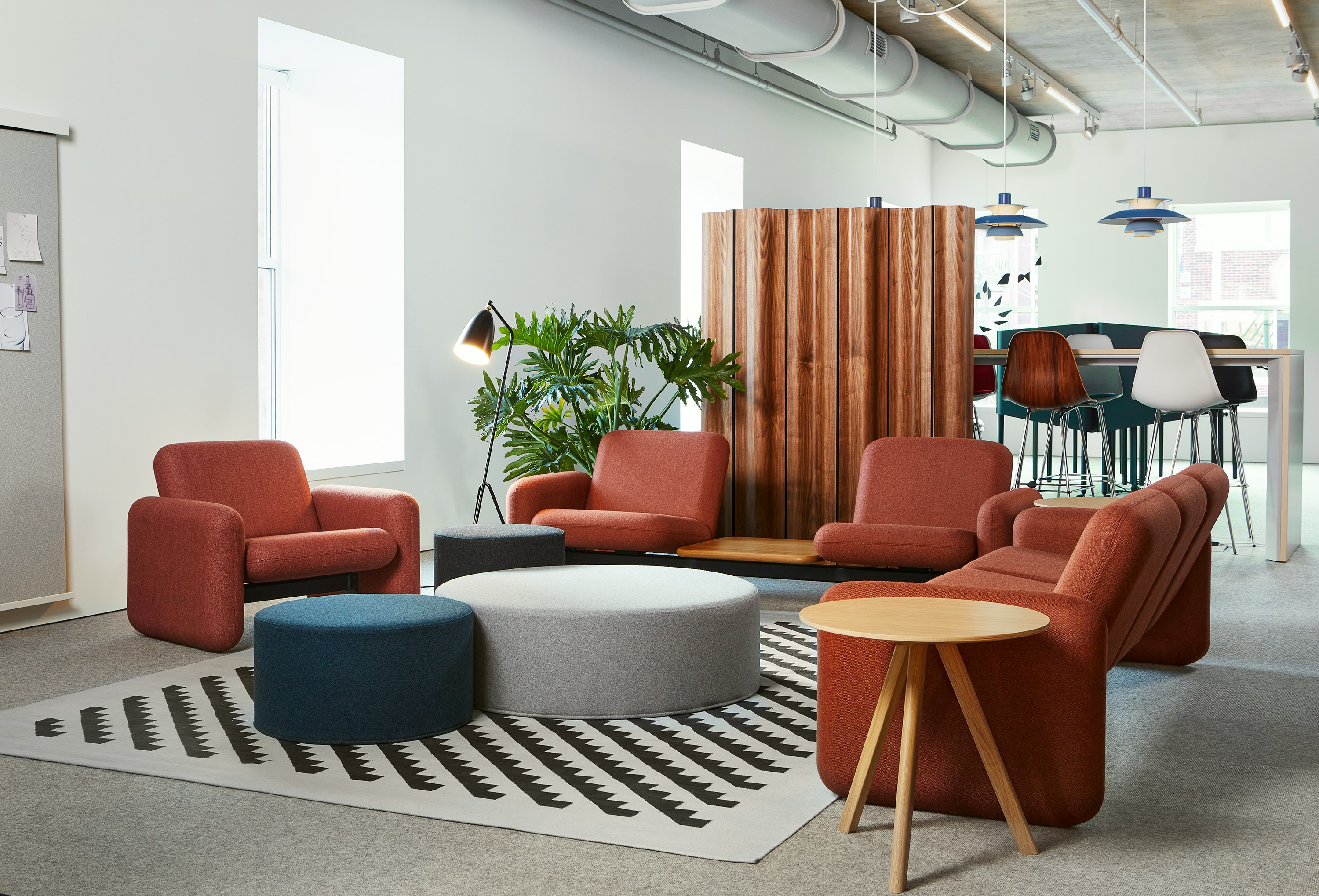 A Wilkes Modular Sofa Group Chair, Two Seat Sofa with white oak table and Three Seat Sofa all in orange-brown surround three poufs in blue and grey and a patterned rug. Behind is a Headway Table and Eames Stools.