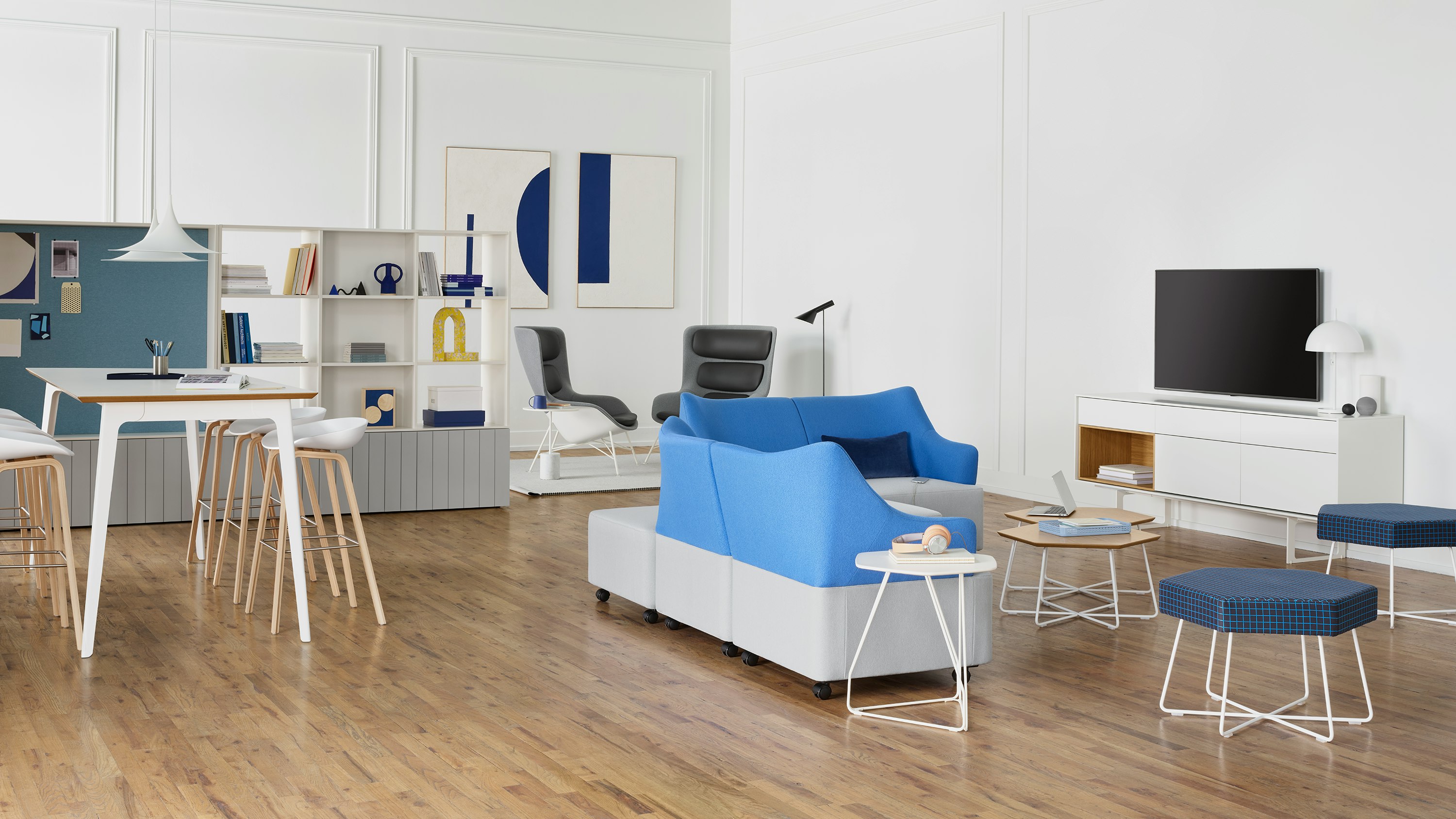 Light grey and blue Plex Lounge Furniture around two Pollen Tables in oak with a white metal base, two Pollen Stool in a blue patterned textile with a white base, and a white Polygon Wire Table. The scene is set in front of a collaborative space and another lounge area. The room has white walls.