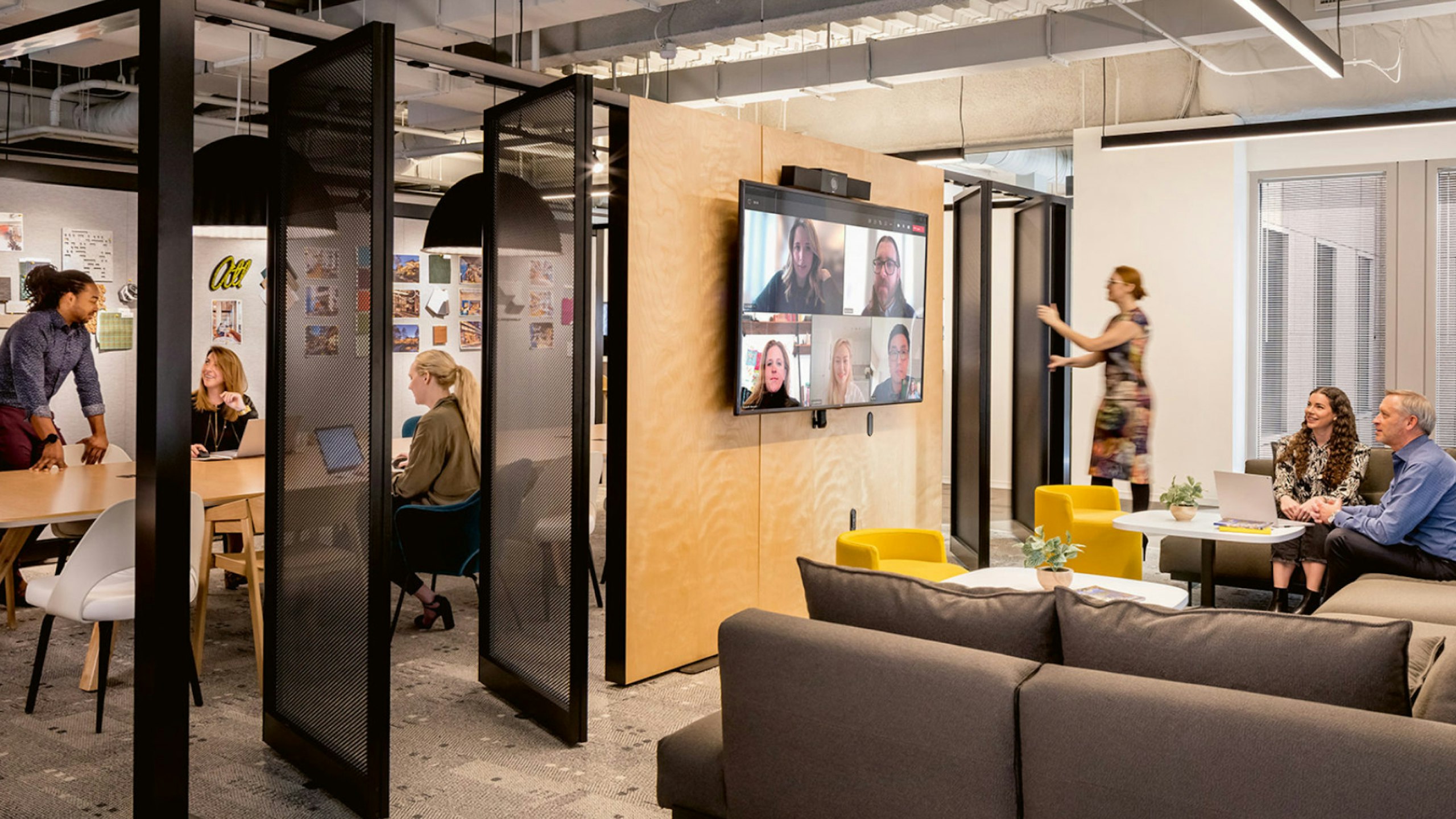 Two employees participating in a video meeting with their colleagues while three other employees interact in a private space.