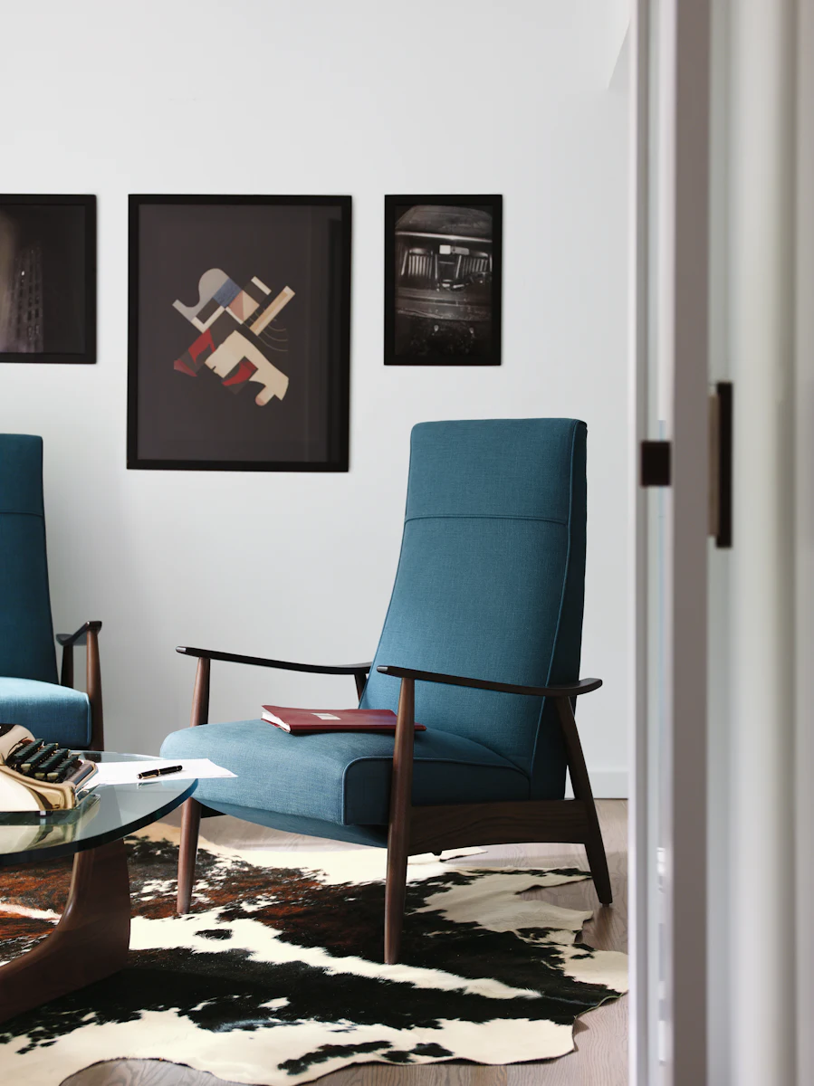 Wall with multiple framed art pieces in a modern living space; blue upholstered chairs with wooden arms.