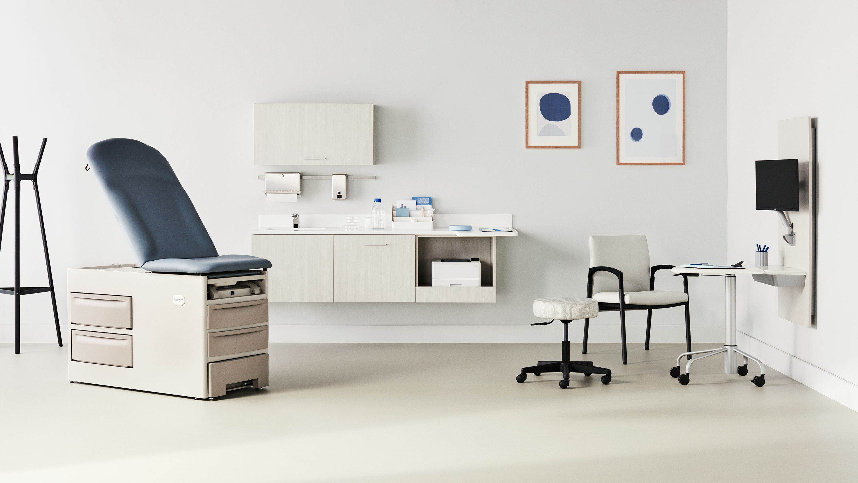 Exam room with a Brewer Company exam table in a soft white base and a blue upholstered back and seat with Mora casework in a light wood laminate on the back wall and Intent solution wall in a light wood laminate to the side that includes a mobile, height-adjustable table and is surrounded by a Valor side chair in a light gray upholstery and black base and a physician stool.