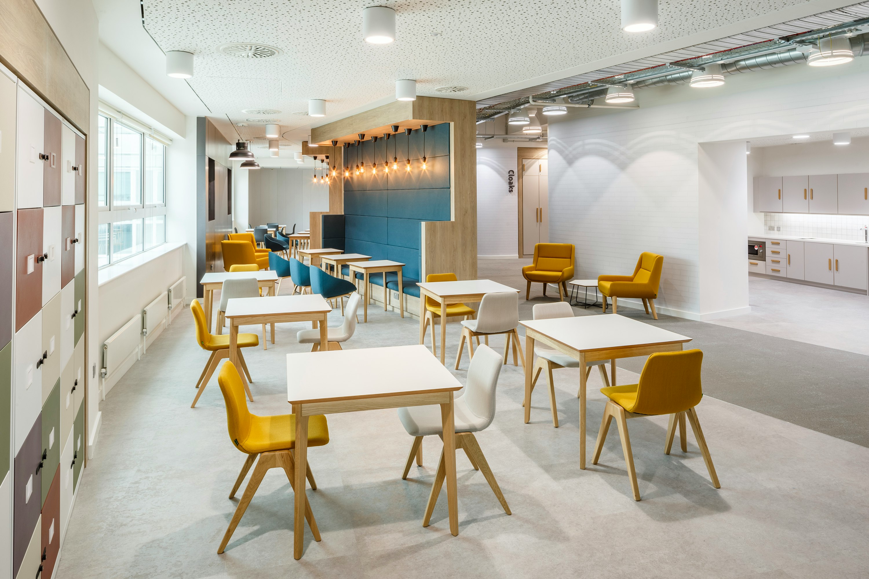 A yellow and a white naughtone Viv Wood Chair stationed at a square Dalby Café Table. Multiple arrangements of this fill an empty café.