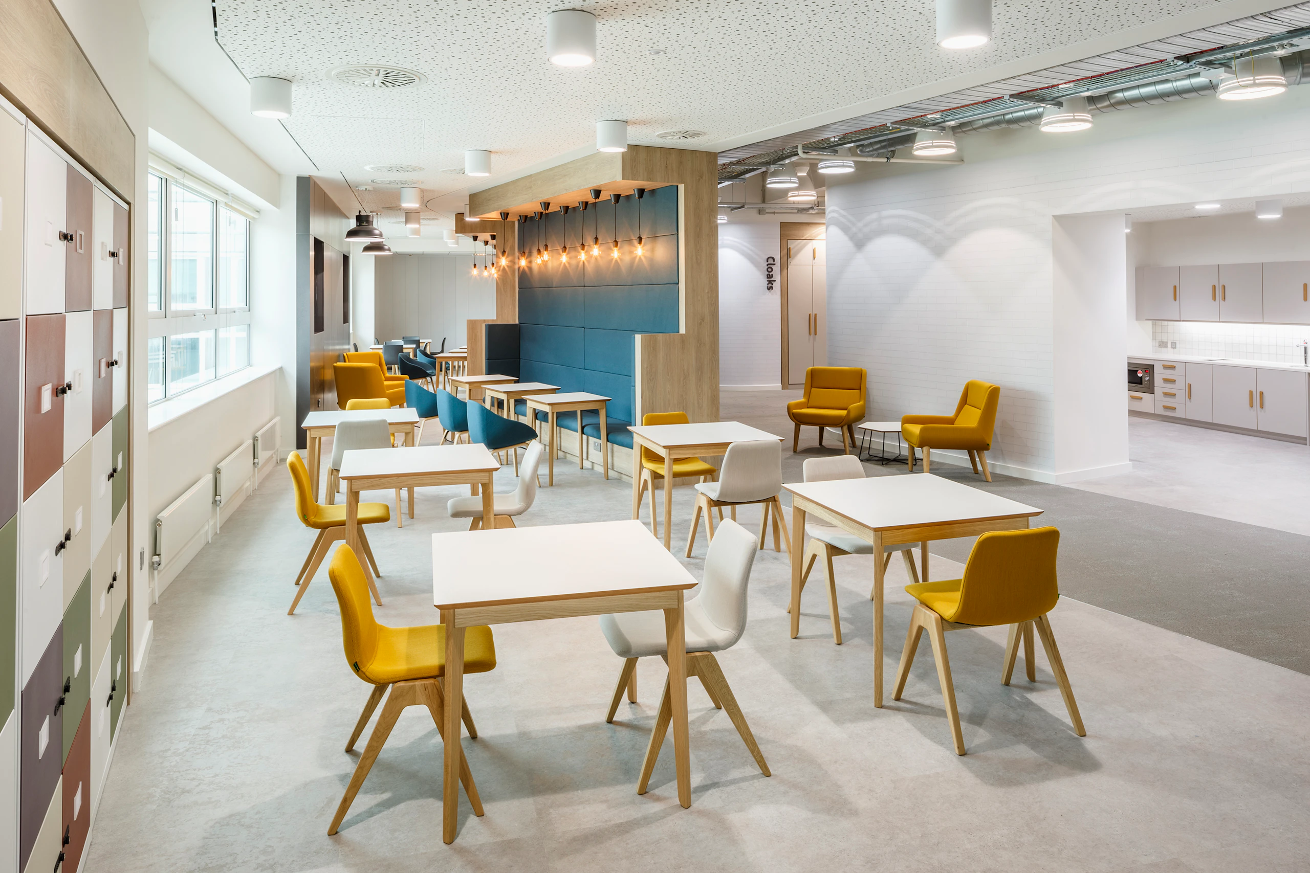 A yellow and a white naughtone Viv Wood Chair stationed at a square Dalby Café Table. Multiple arrangements of this fill an empty café.