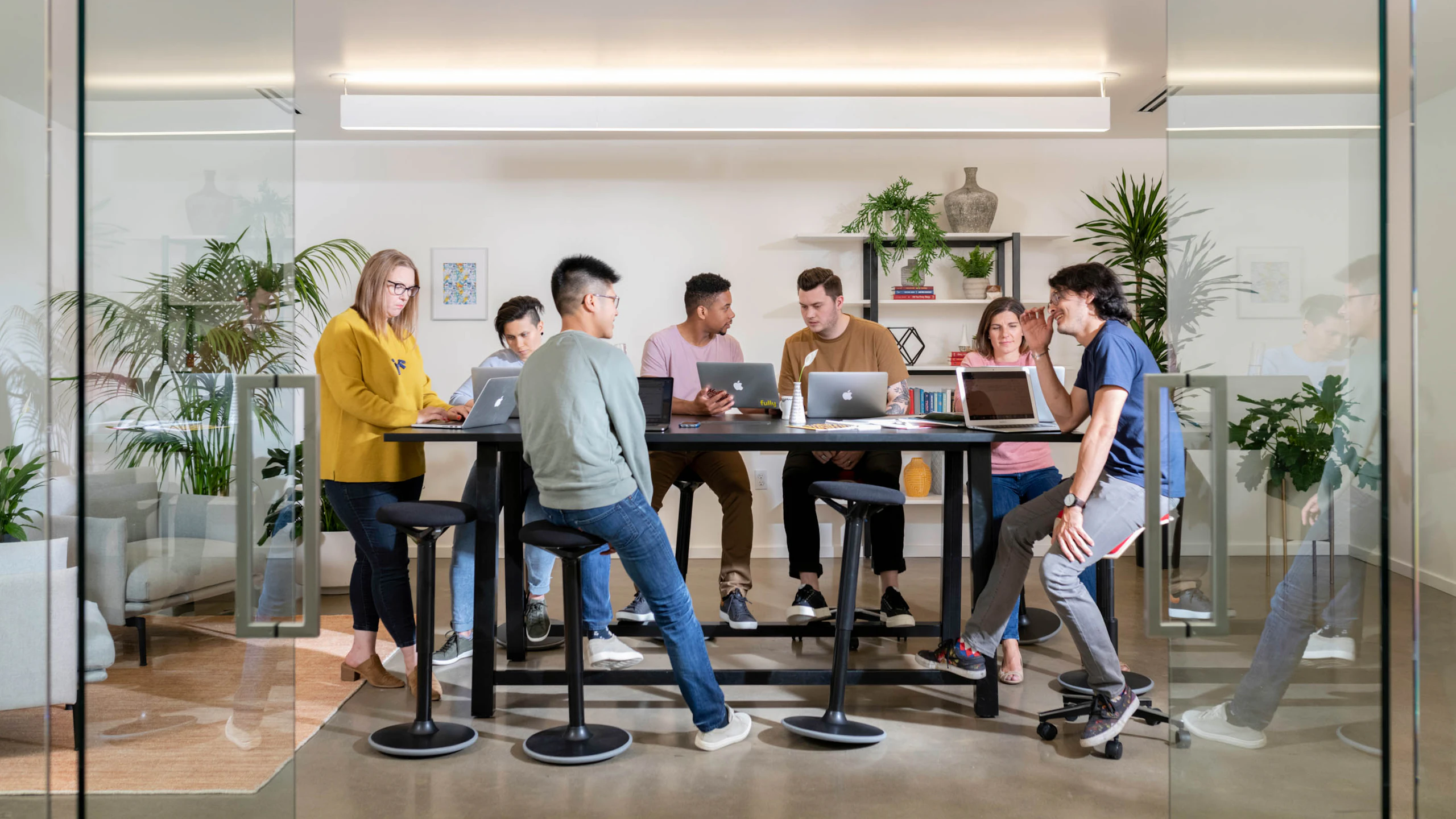 Seven people sit around a black bar-height table on black stools in a conference room with glass walls.