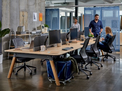 A long conference table in a light woodgrain surrounded by black ReGeneration Chairs by Knoll. At the end of the table two people meet by a monitor.
