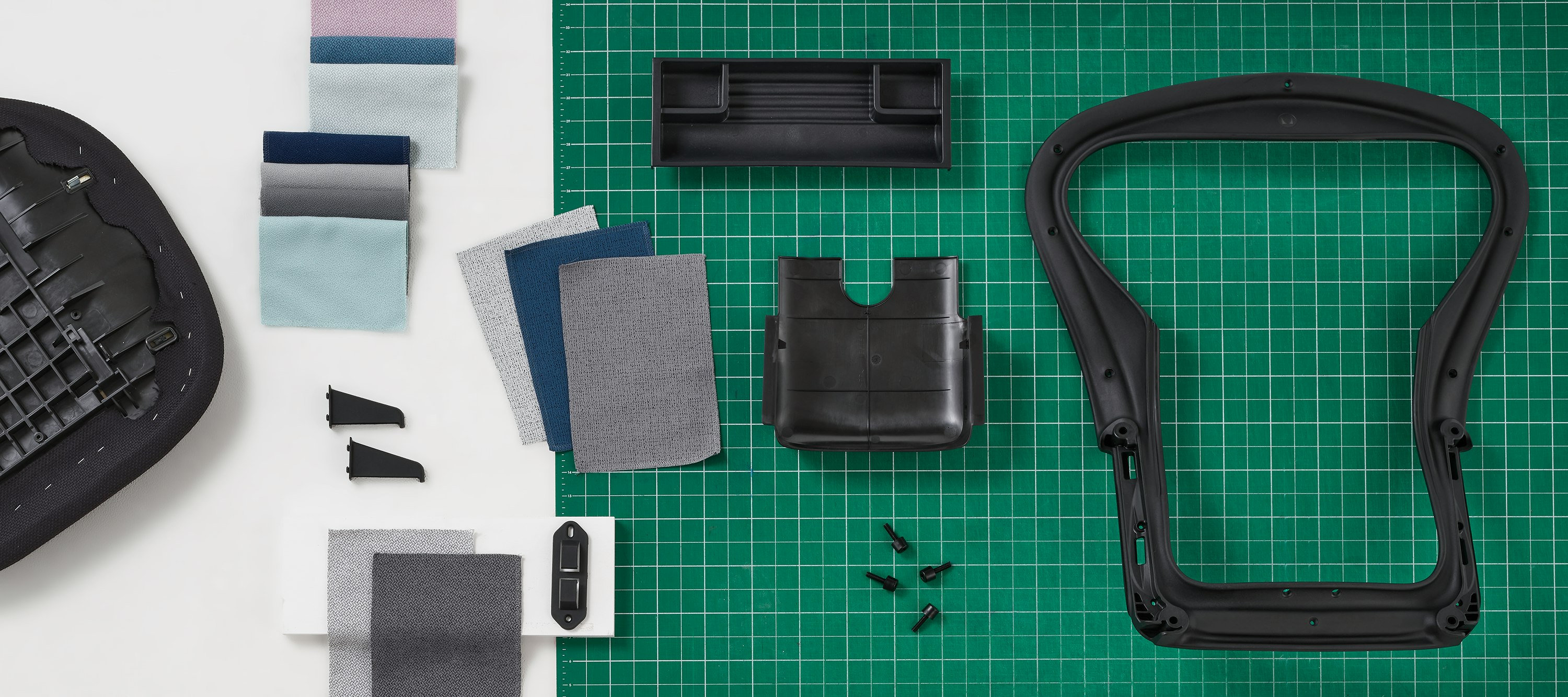 Overhead view of a black Aeron back frame and Aeron Tilt cover, a line of both folded and fanned Revenio textile color swatches, a Sayl seat pan and OE1 Trolly ocean bound plastic clips arranged across a half white and half green table top.