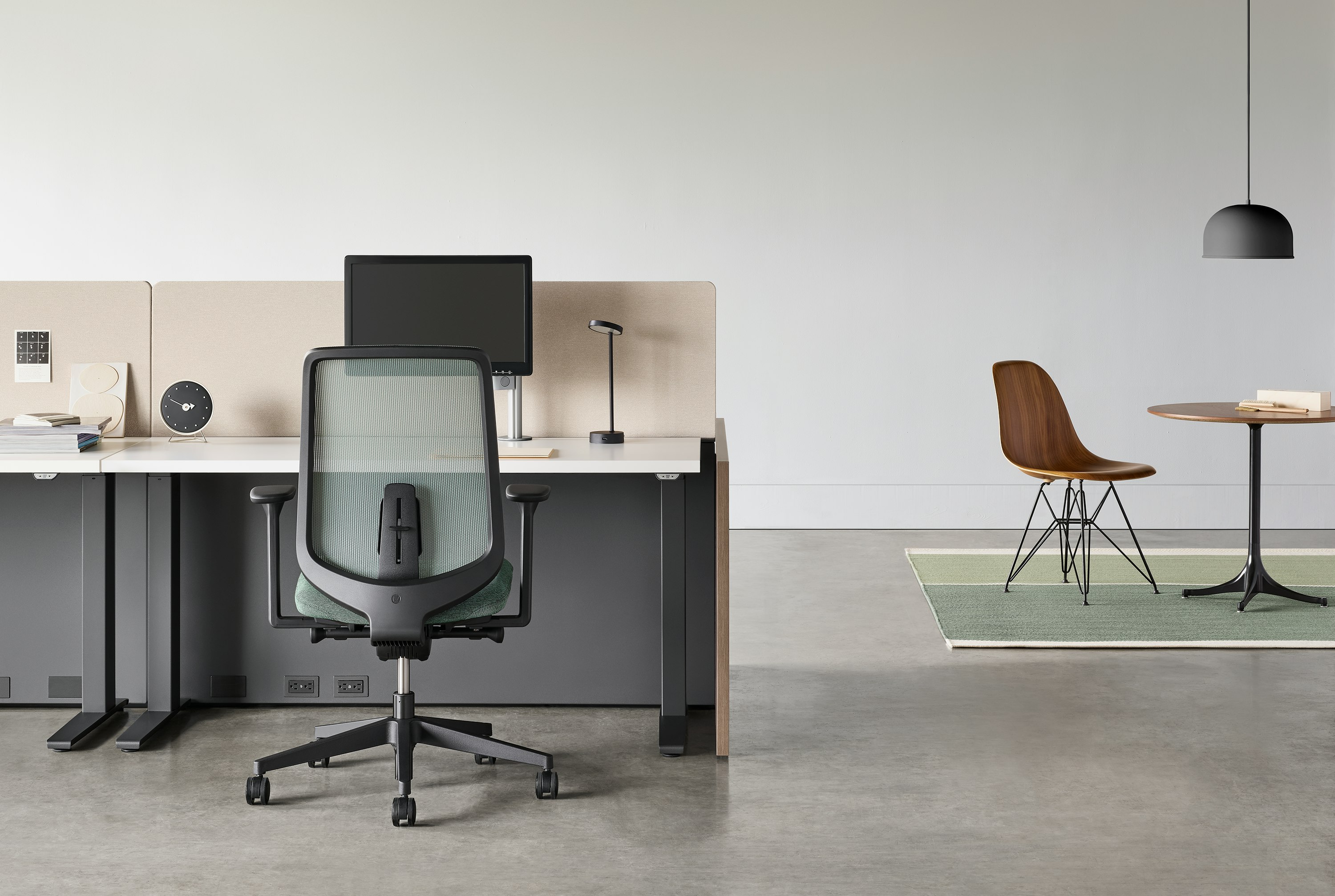 A black Verus chair with a black Motia Sit-to-Stand Table and Canvas Channel workstation with a side table and chair in the background.