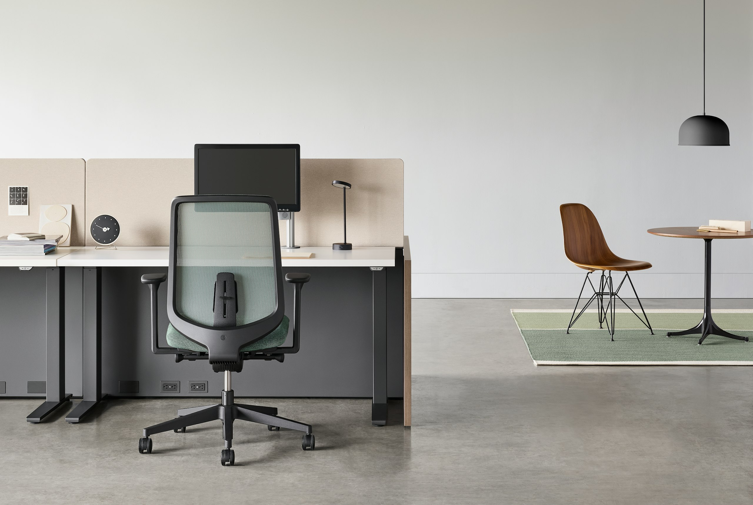 A black Verus chair with a black Motia Sit-to-Stand Table and Canvas Channel workstation with a side table and chair in the background.