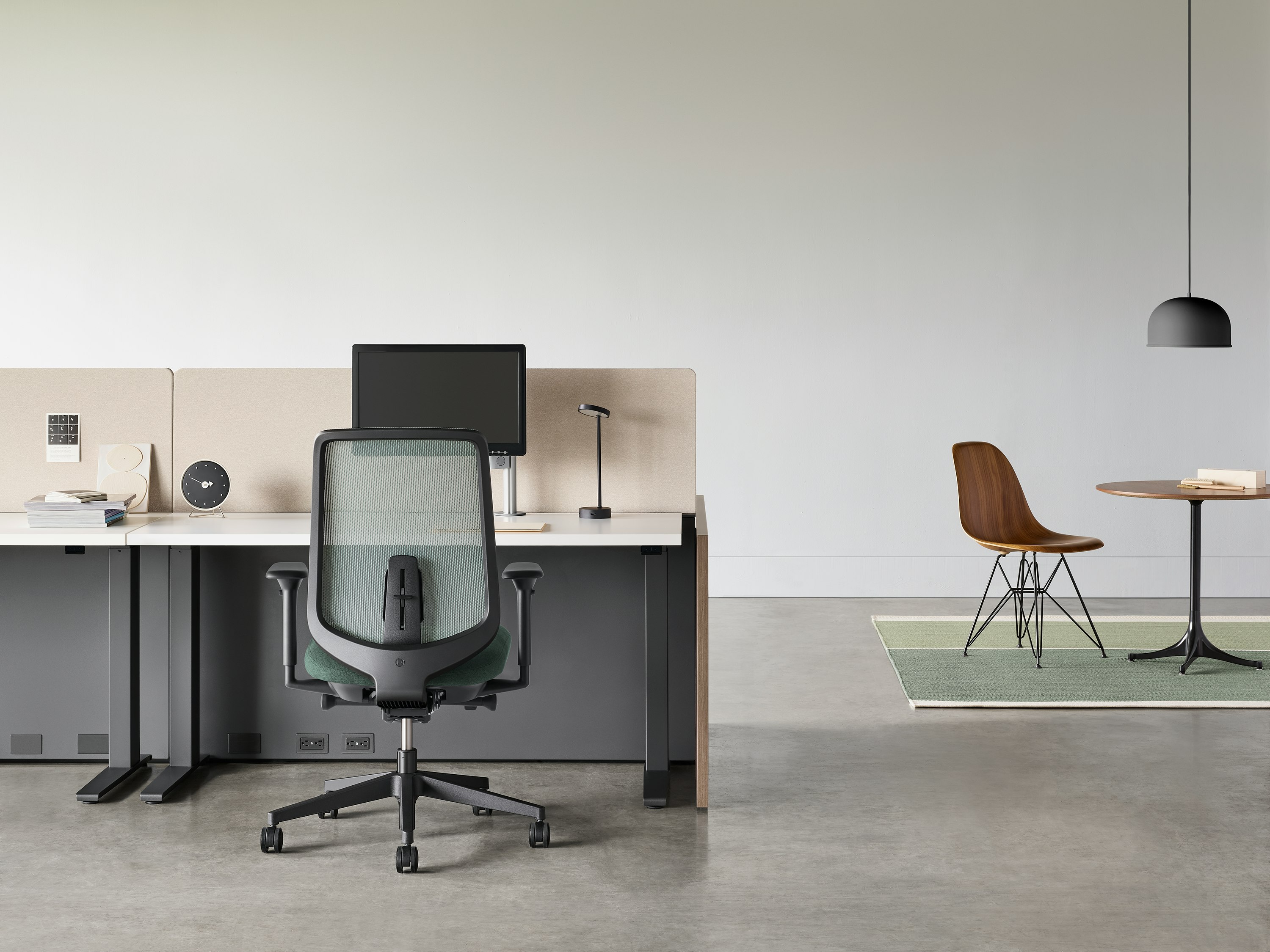 A black Verus chair with a black Motia Sit-to-Stand Table and Canvas Channel workstation with a Nelson Pedestal Table and Eames Molded Wood Side Chair in the background.