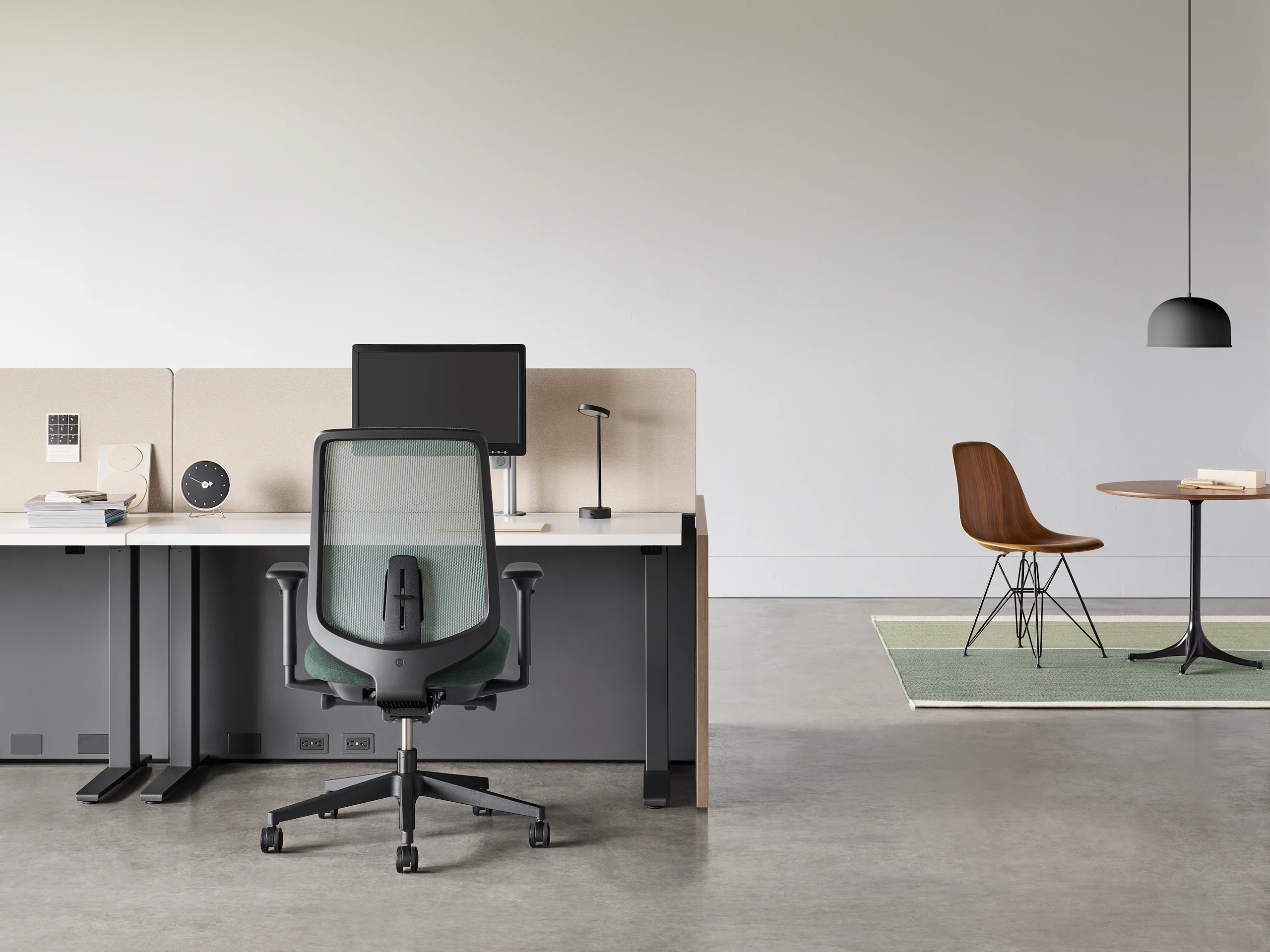 A black Verus chair with a black Motia Sit-to-Stand Table and Canvas Channel workstation with a Nelson Pedestal Table and Eames Molded Wood Side Chair in the background.