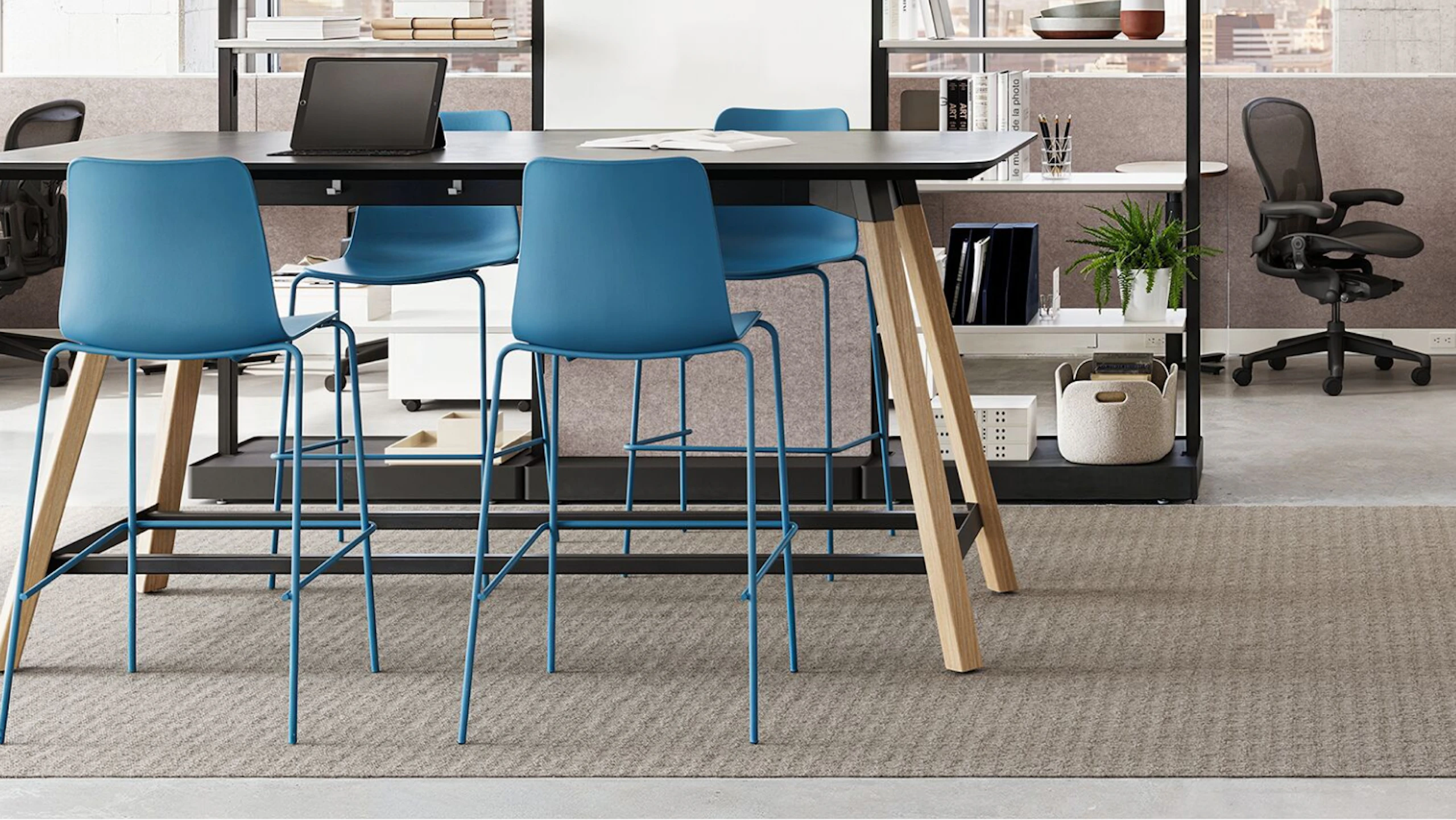 A black NaughtOne Morse Bar Table with wooden legs surrounded by four blue bar-height chairs in a team neighborhood setting designed to facilitate change, reduce disruption, and improve resiliency.