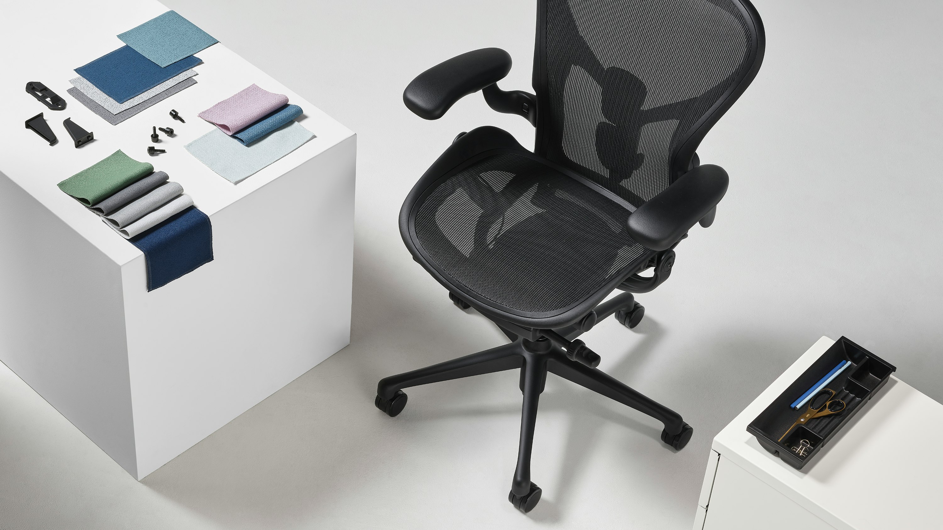 A black Aeron chair viewed from above, is positioned next to both a white cube, on top of which sits a folded arrangement of Revenio textile swatches and OE1 Trolly plastic clips, and a Tu Storage Pedestal with the top drawer open revealing a black Utility Tray made of ocean-bound plastic.