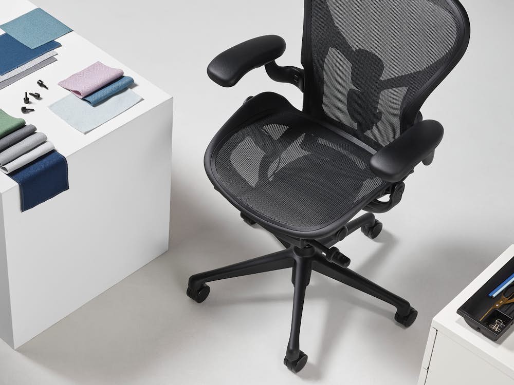 A black Aeron chair viewed from above, is positioned next to both a white cube, on top of which sits a folded arrangement of Revenio textile swatches and OE1 Trolly plastic clips, and a Tu Storage Pedestal with the top drawer open revealing a black Utility Tray made of ocean-bound plastic.
