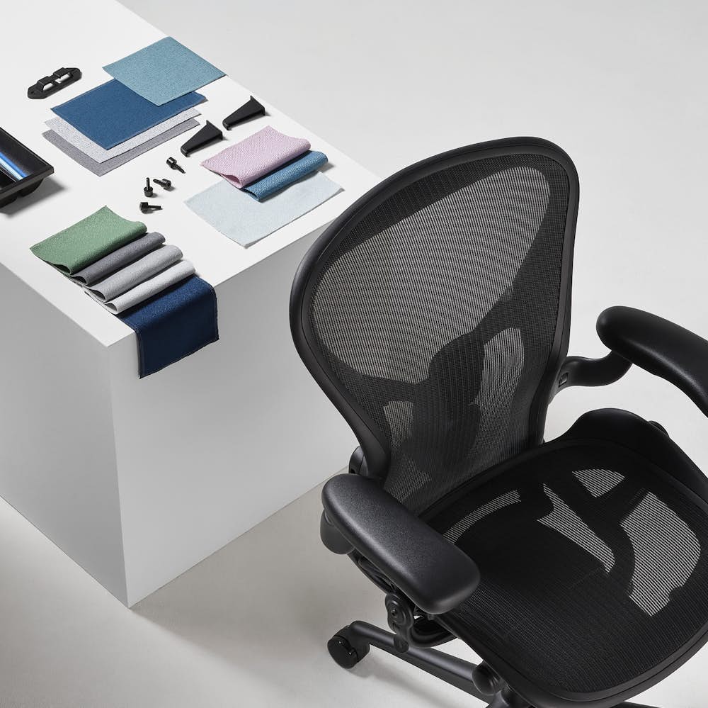 A black Aeron and Sayl office chair viewed from above, are positioned next to a white display cube, on top of which an arrangement of folded Revenio textile swatches, OE1 trolly plastic clips, and a Tu Pedestal Utility Tray made from ocean-bound plastics are all displayed.