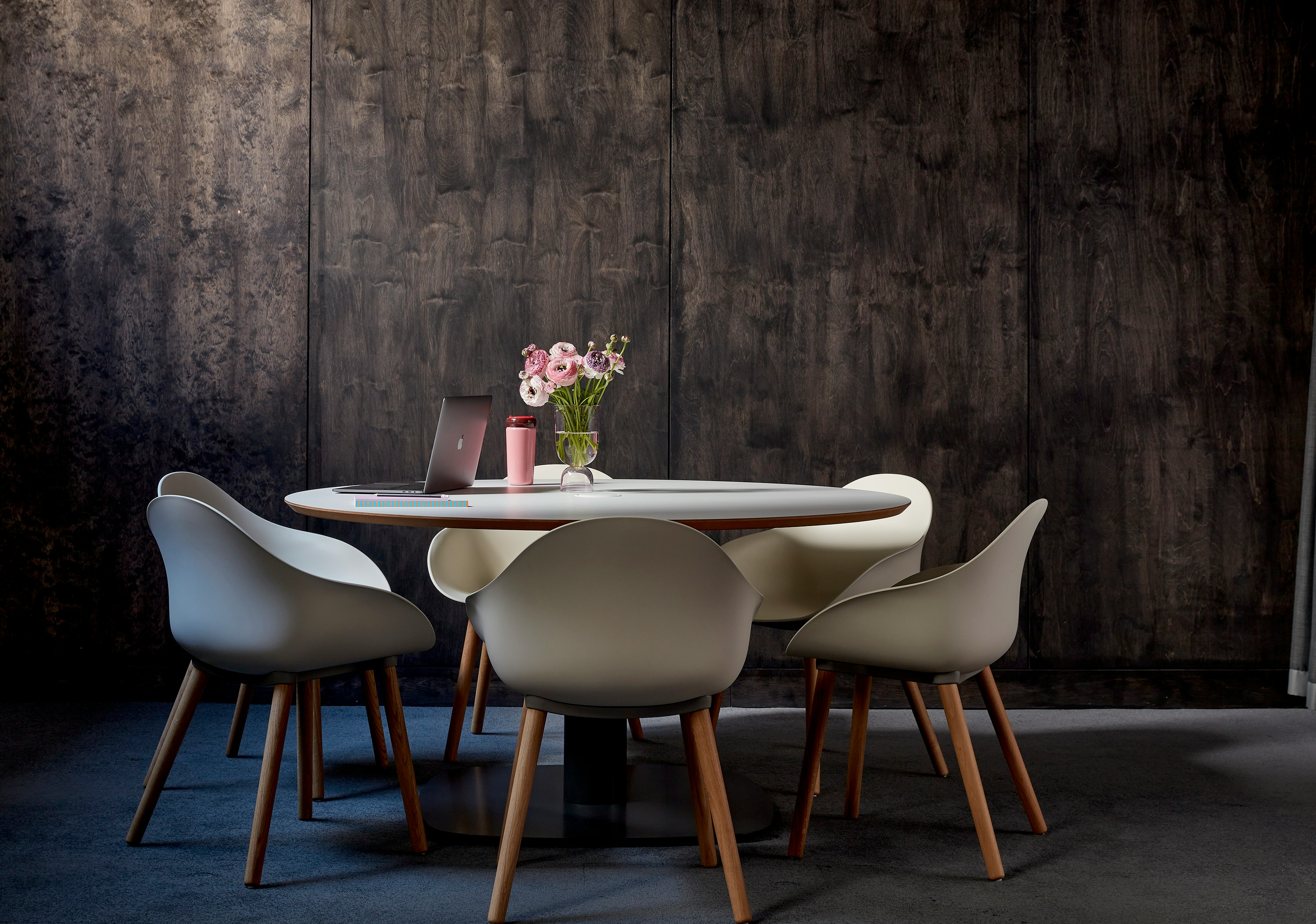 A round Megaped table surrounded by Oyster white Ruby Side chairs with oak dowel bases, set against a wooden wall.