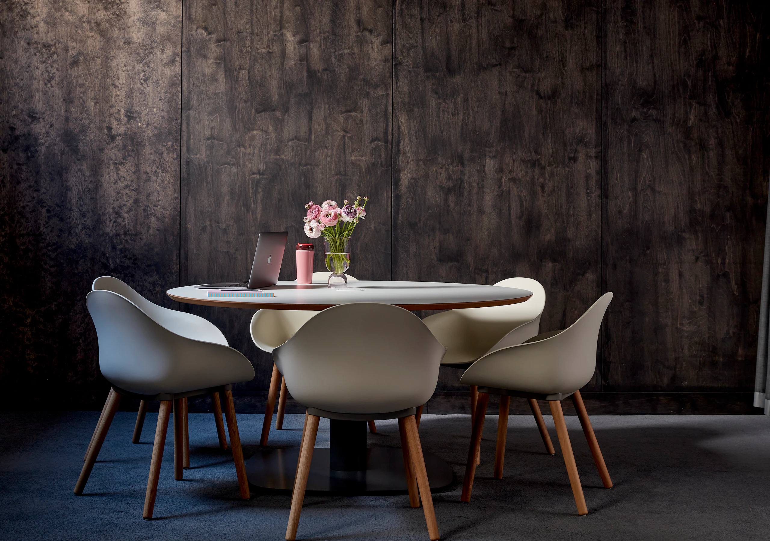 A round Megaped table surrounded by Oyster white Ruby Side chairs with oak dowel bases, set against a wooden wall.