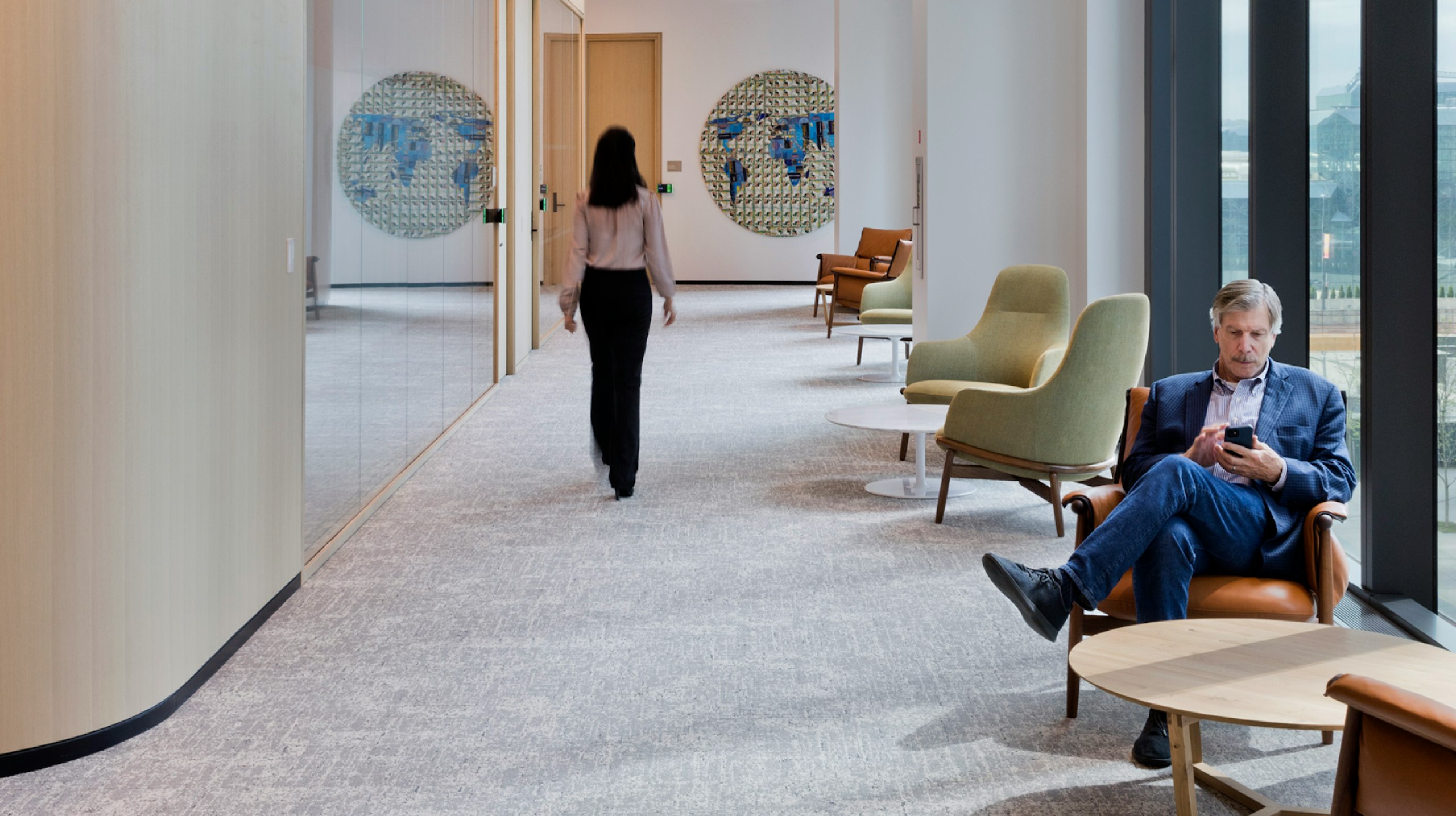 An individual in blue seated next to a window using their phone while a person walks past several seating areas at the BlackRock offices in New York City