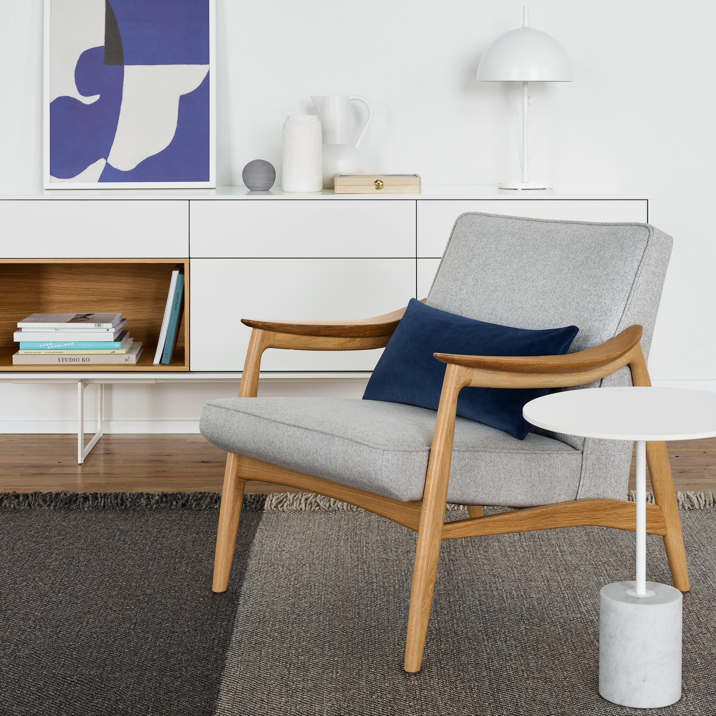 An Aspen Lounge Chair in a light gray textile next to a white Jay Table. The scene is set on top of a gray rug in front of a white Aura Credenza with various decorative elements against a white wall.