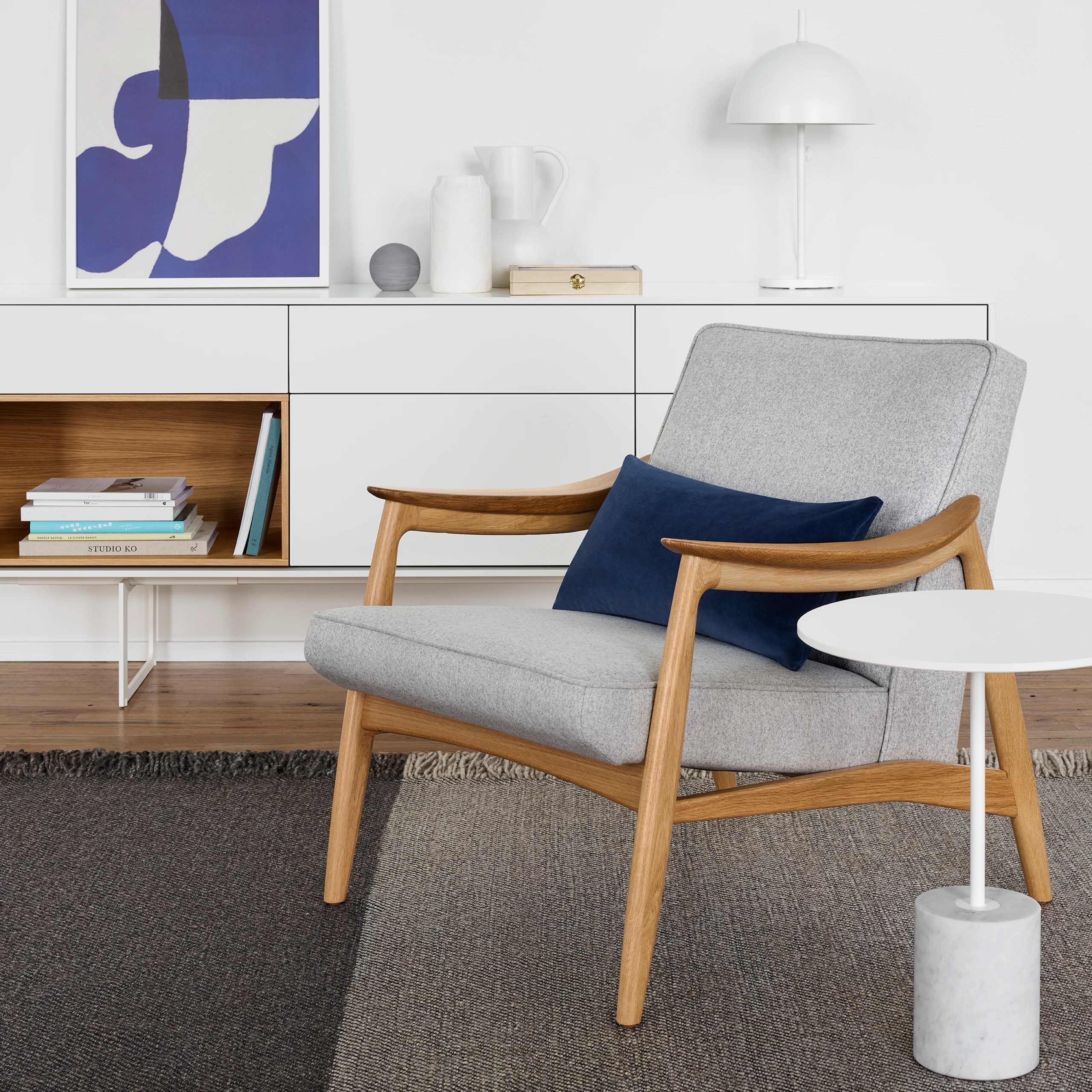 An Aspen Lounge Chair in a light gray textile next to a white Jay Table. The scene is set on top of a gray rug in front of a white Aura Credenza with various decorative elements against a white wall.