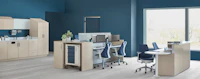 An Ethospace nurse station in a light wood finish, consisting of a reception desk, two-sided workstation for sitting or standing, Mora casework in matching wood, and a supply cart with blue drawers.
